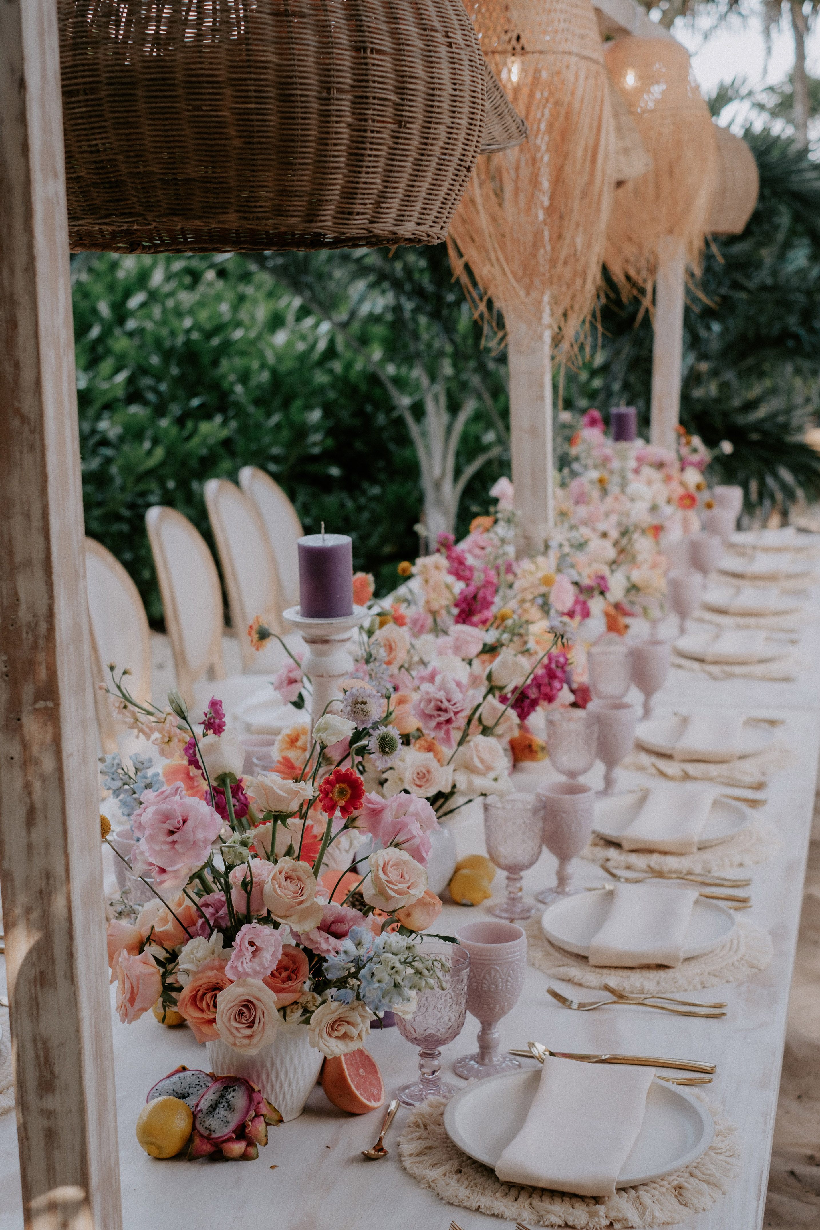 Vibrant floral outdoor dining table with purple glasses, macrame, and tropical fruit.