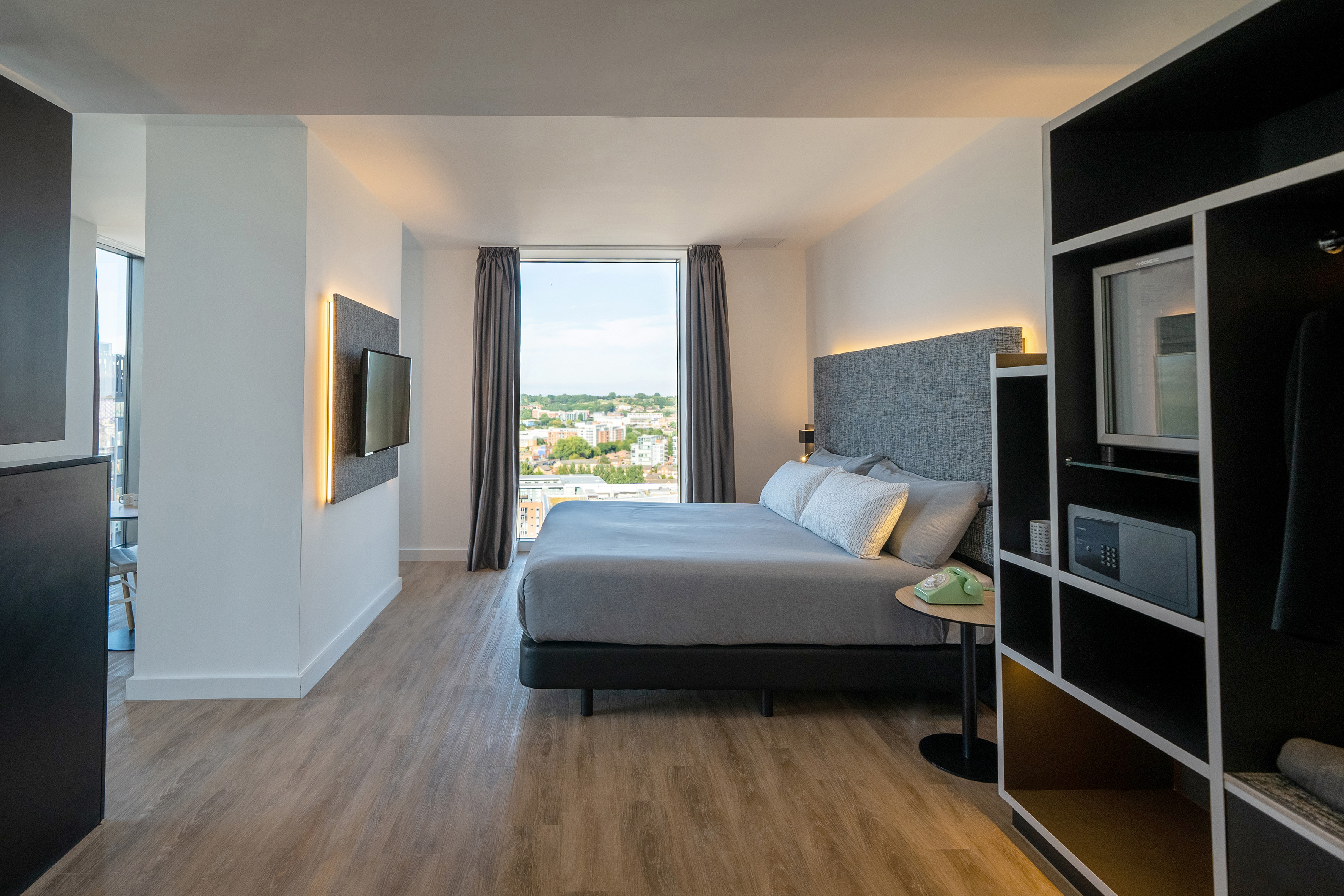Modern hotel room with large grey bed, city view, TV, and integrated storage.