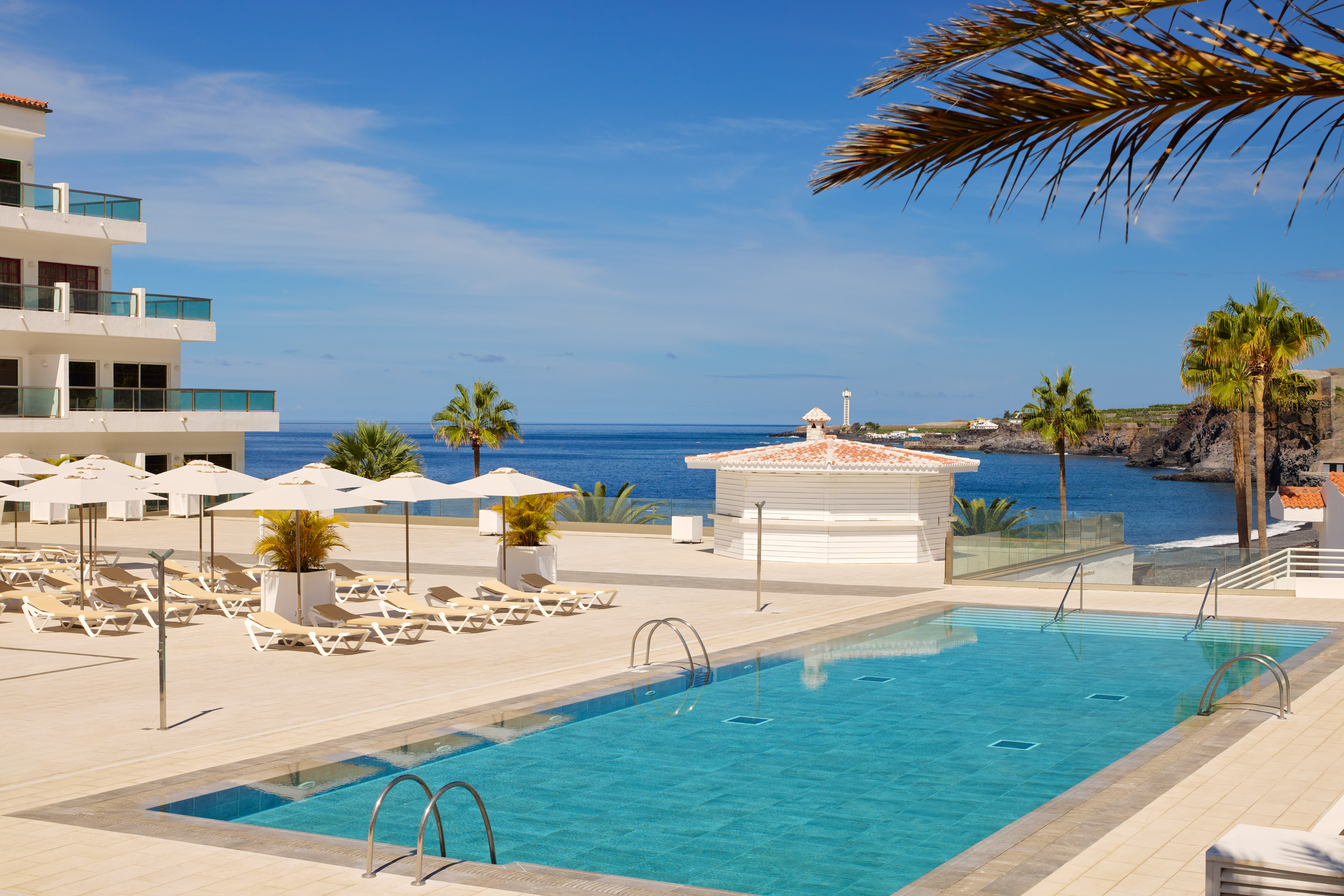 Tropical resort pool, blue ocean view, sun loungers, and palm trees.