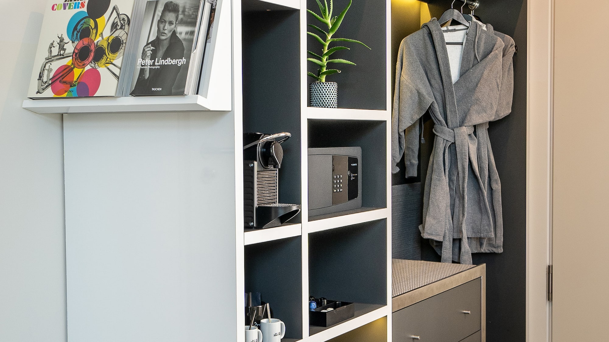 Stylish hotel room wardrobe with Nespresso, safe, bathrobes, slippers, and books.