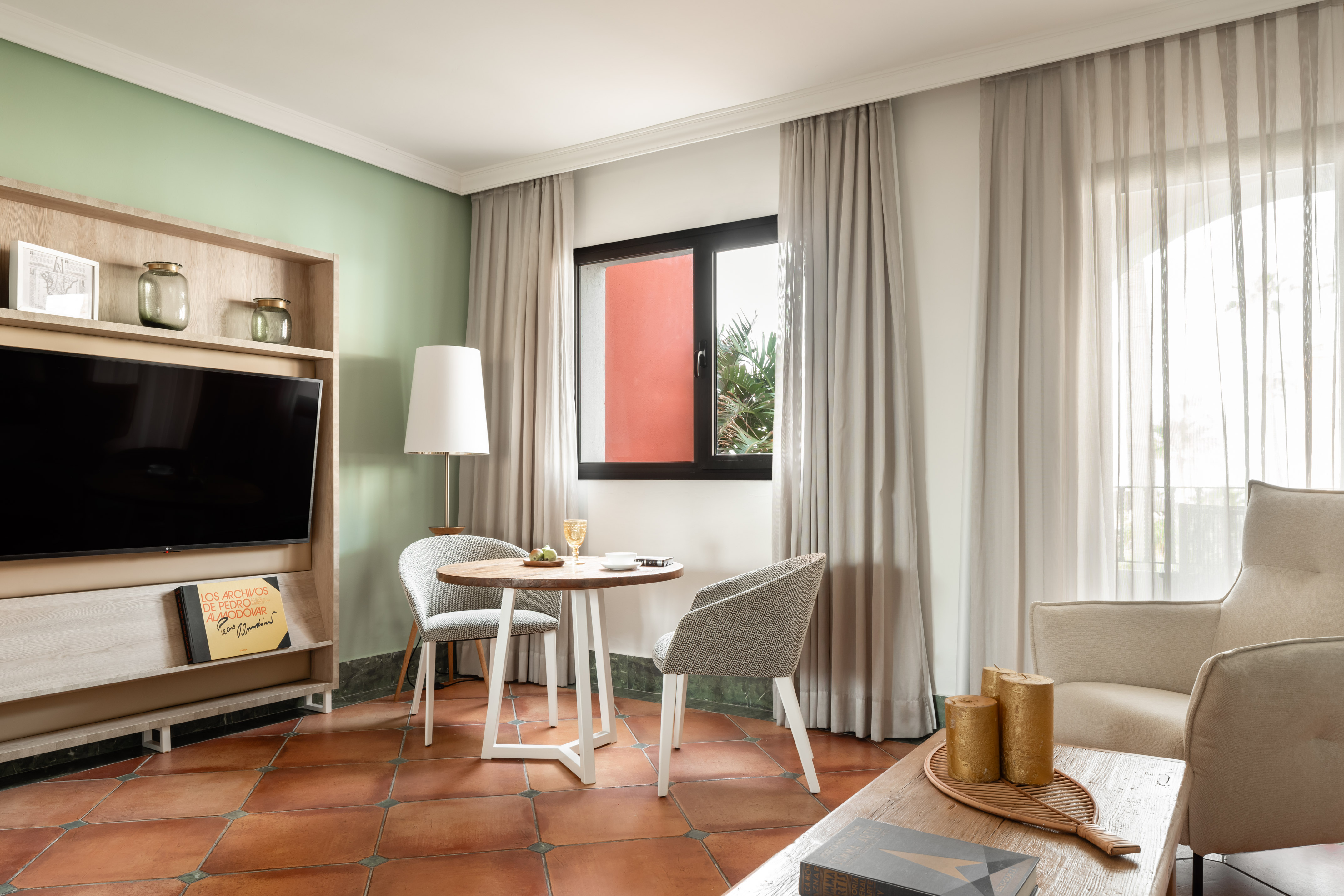 Hotel living room with green wall, TV, dining table, and terracotta floor.