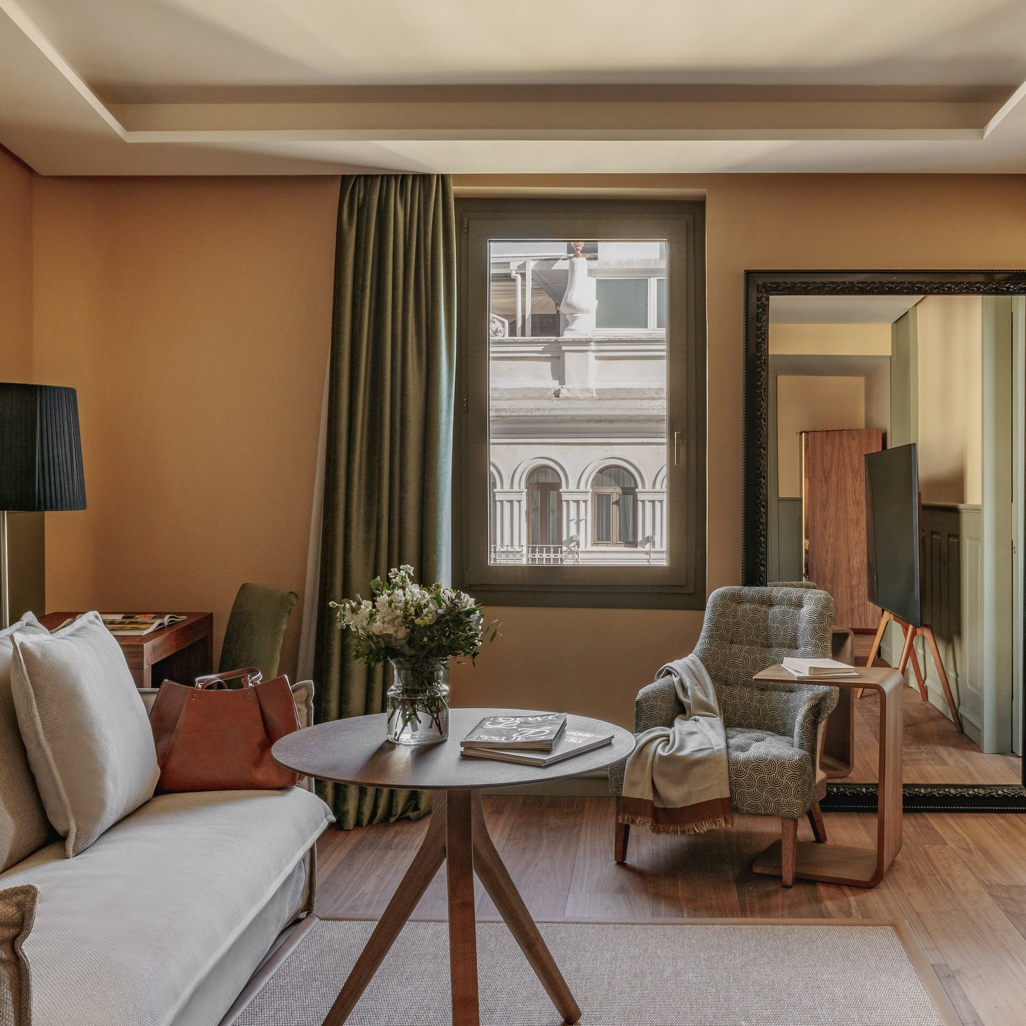 Diseño de suite de hotel con sofá, sillón, mesa, flores y ventana urbana.