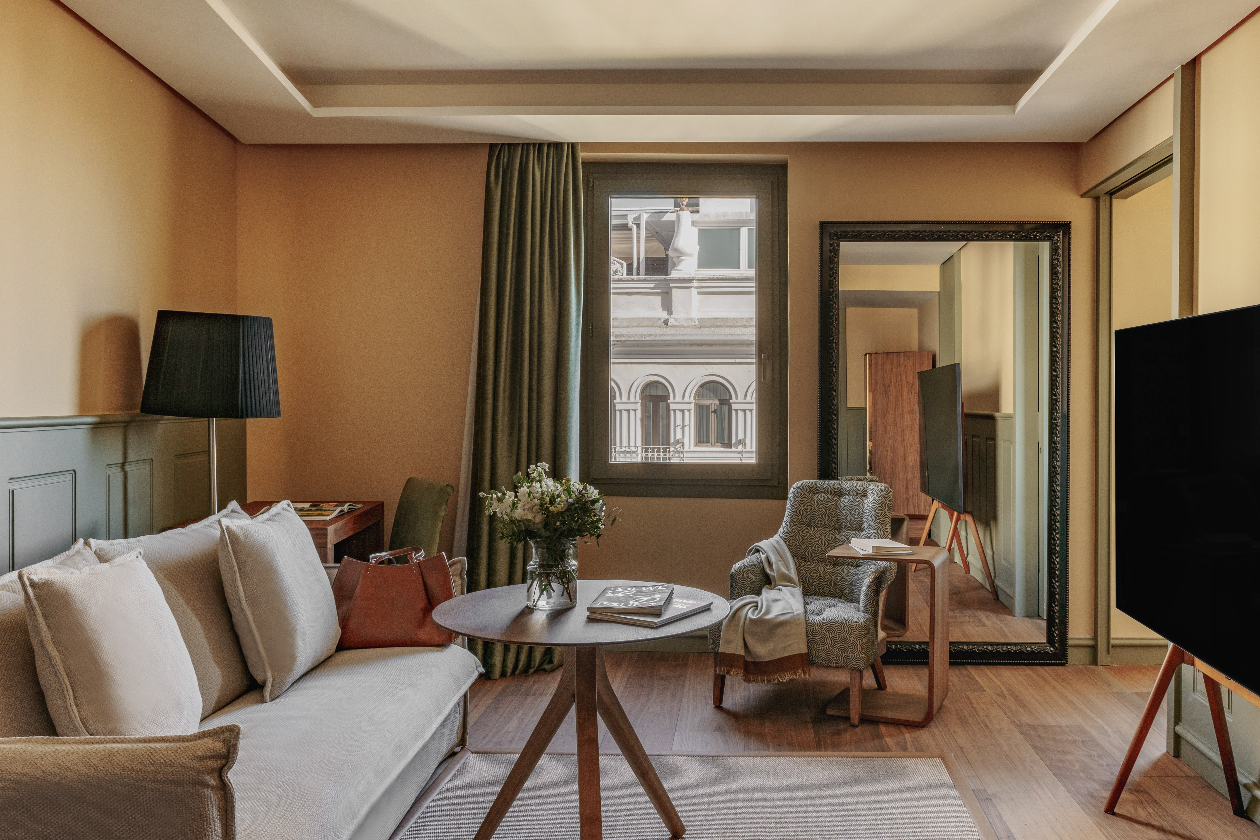 Diseño de suite de hotel con sofá, sillón, mesa, flores y ventana urbana.