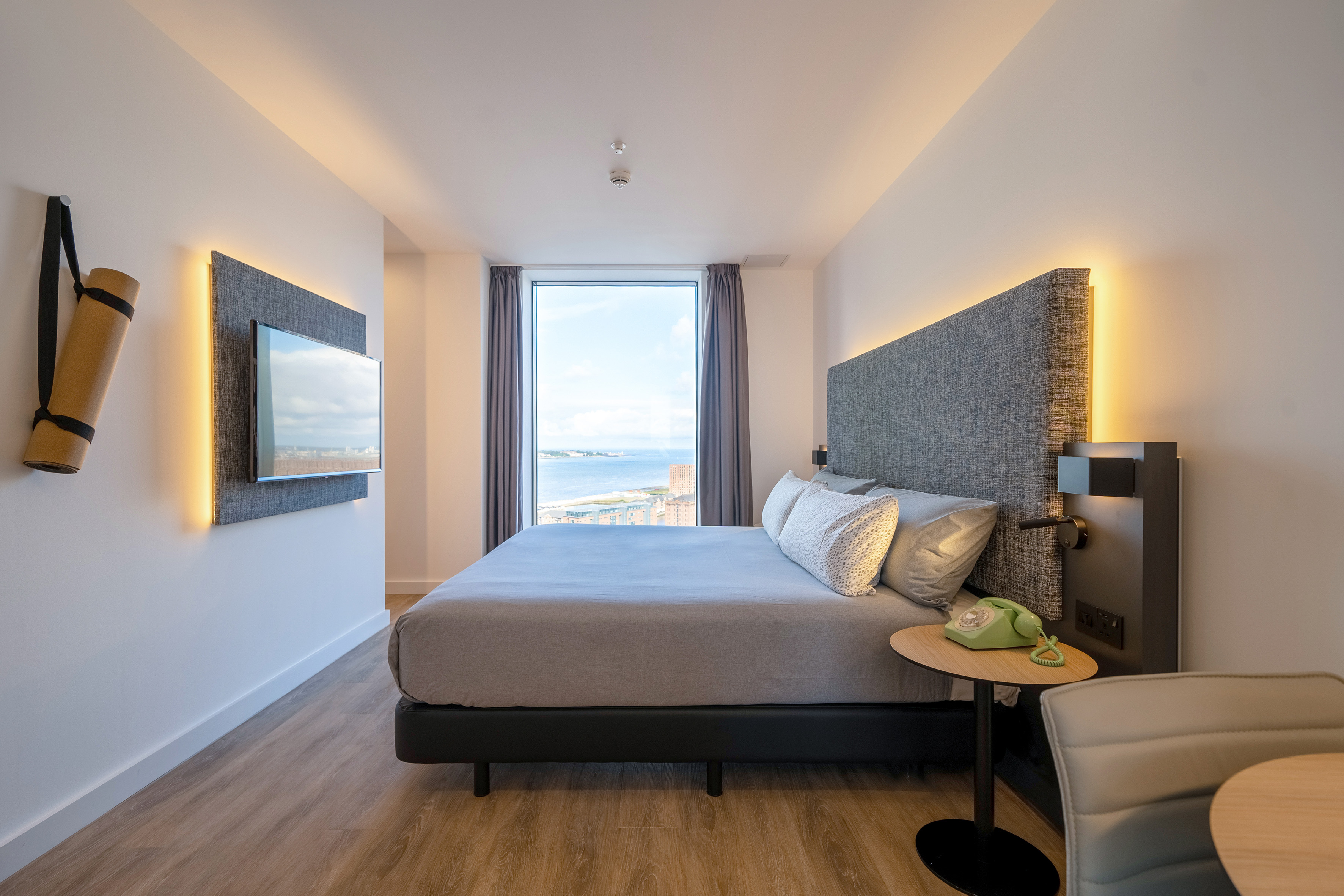 Modern hotel bedroom with sea view, grey bed, wall TV, and yoga mat.