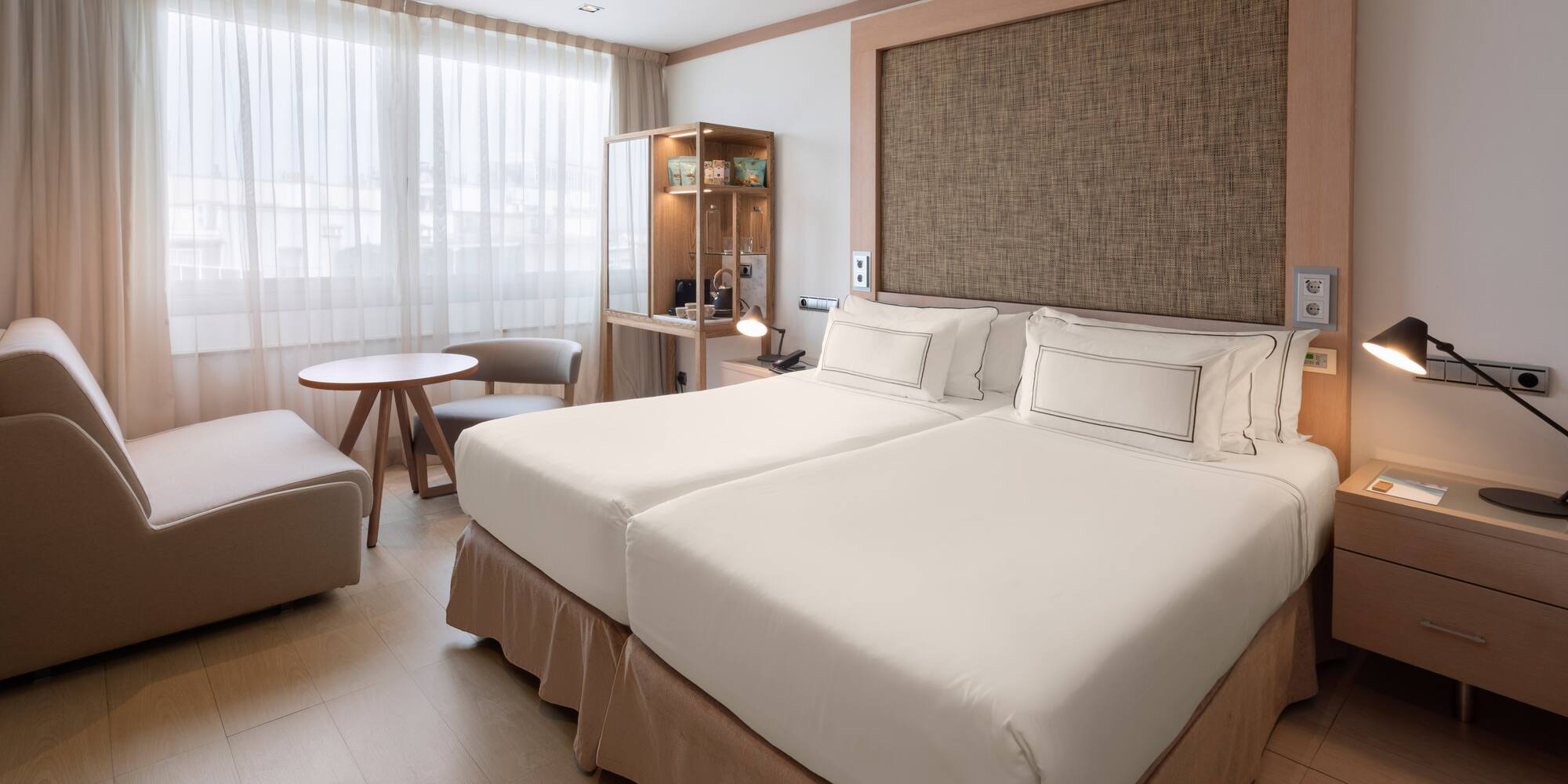 Modern hotel room with twin beds, lounge area, and light wooden floor.