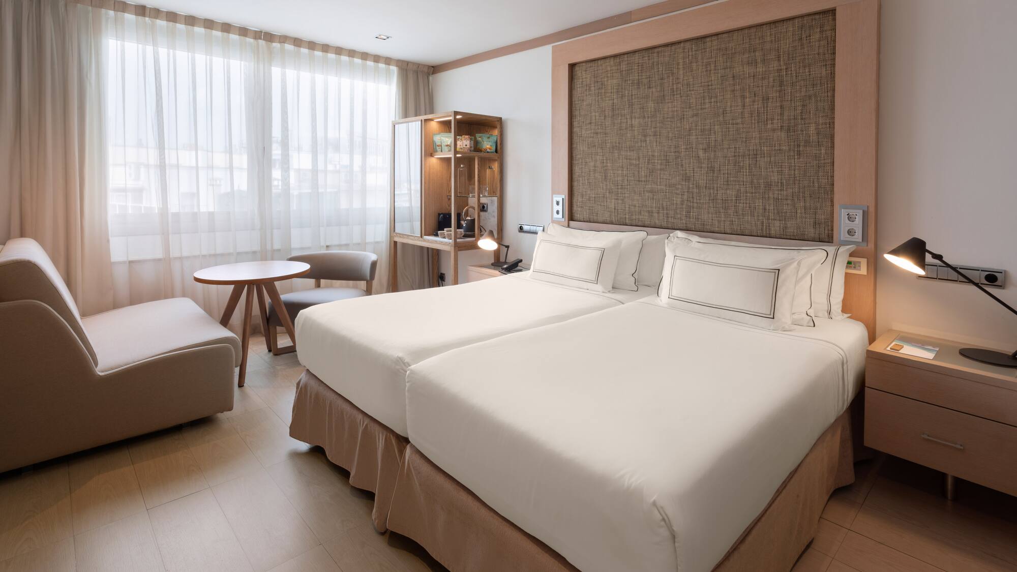 Modern hotel room with twin beds, lounge area, and light wooden floor.