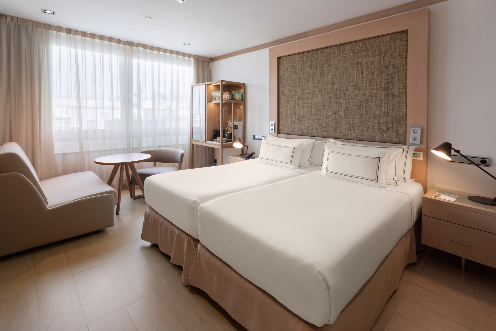 Modern hotel room with twin beds, lounge area, and light wooden floor.
