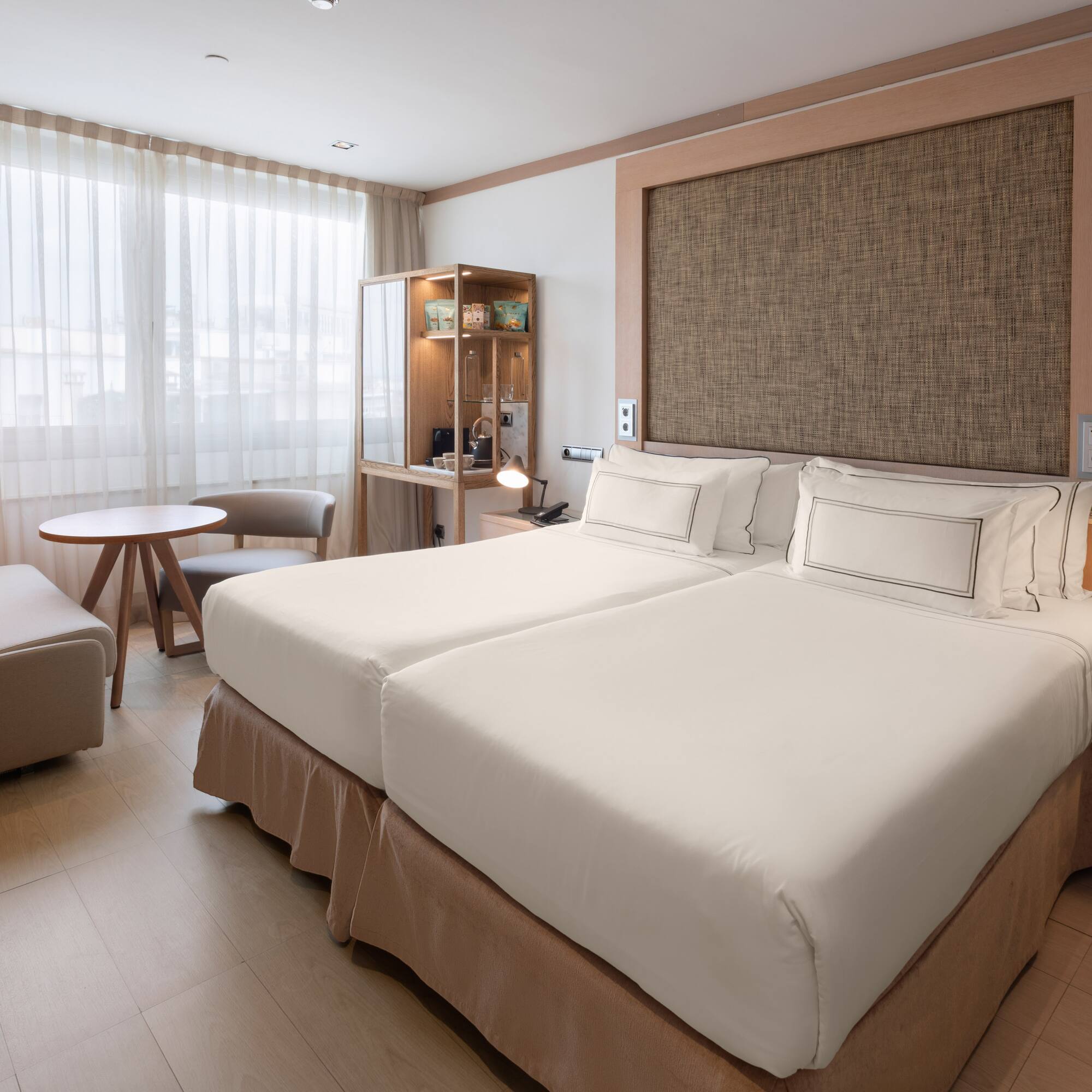 Modern hotel room with twin beds, lounge area, and light wooden floor.