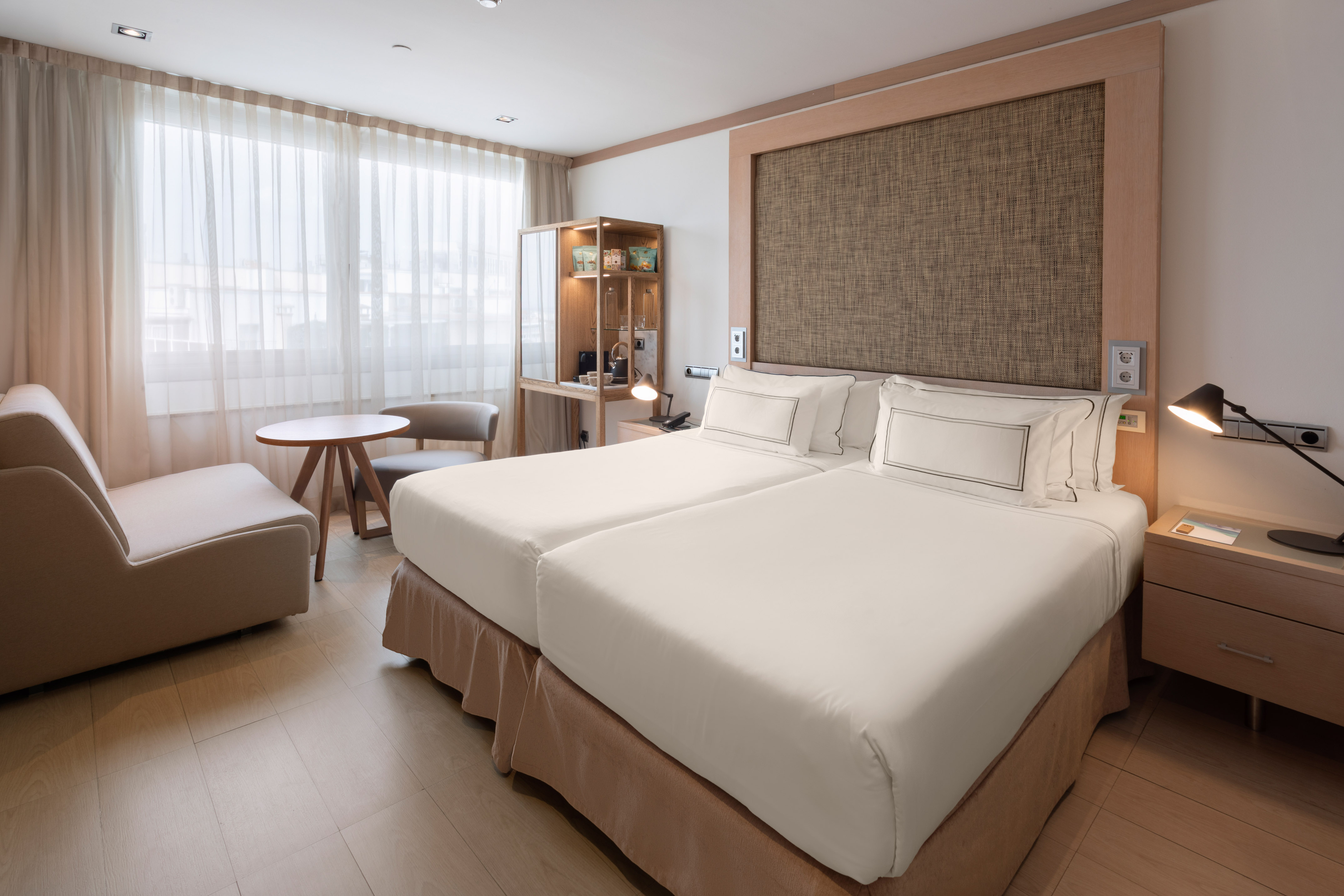 Modern hotel room with twin beds, lounge area, and light wooden floor.