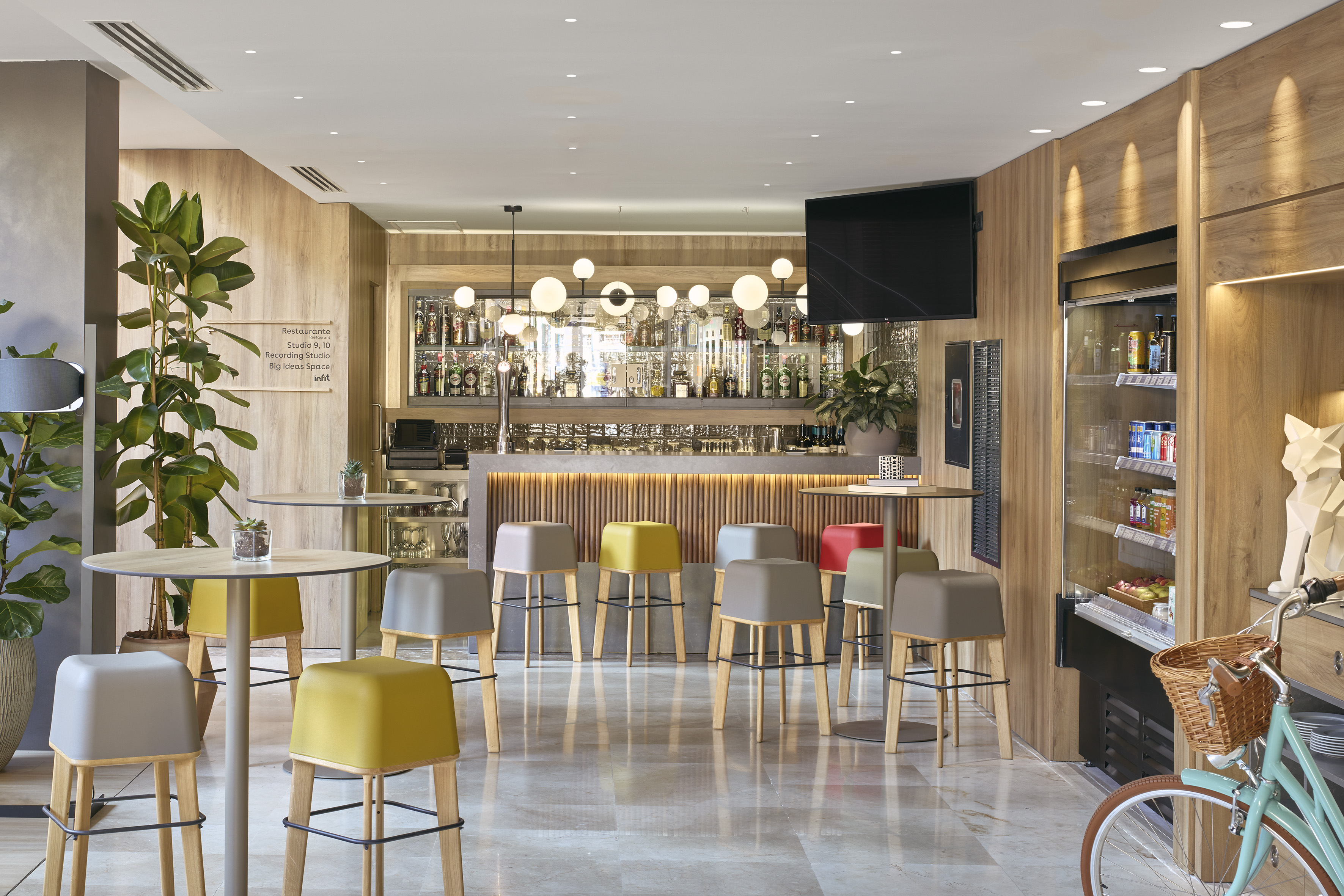 Modern hotel bar with colorful stools, wooden bar, and beverage cooler.