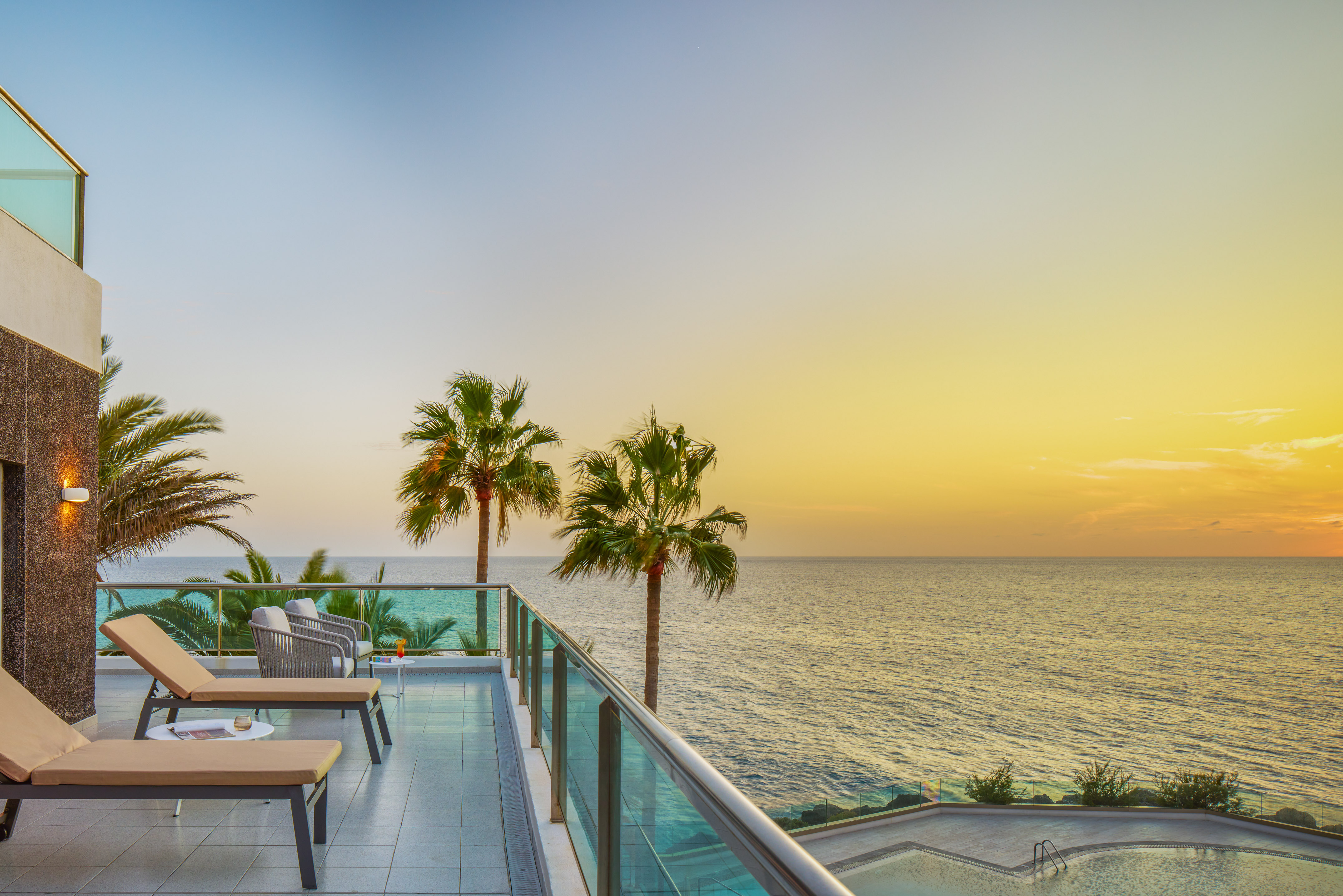 Luxury oceanfront balcony at sunset with lounge chairs and palm trees.