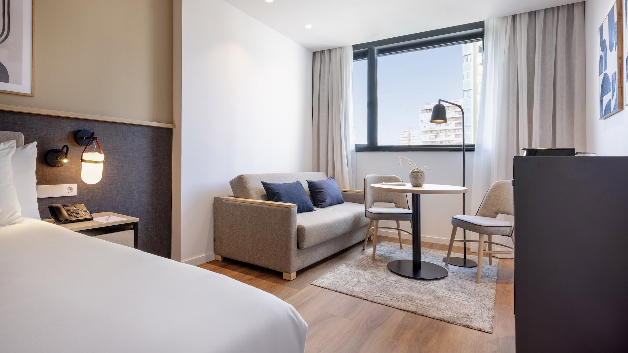 Modern hotel studio with bed, sofa, dining table, and city view window.
