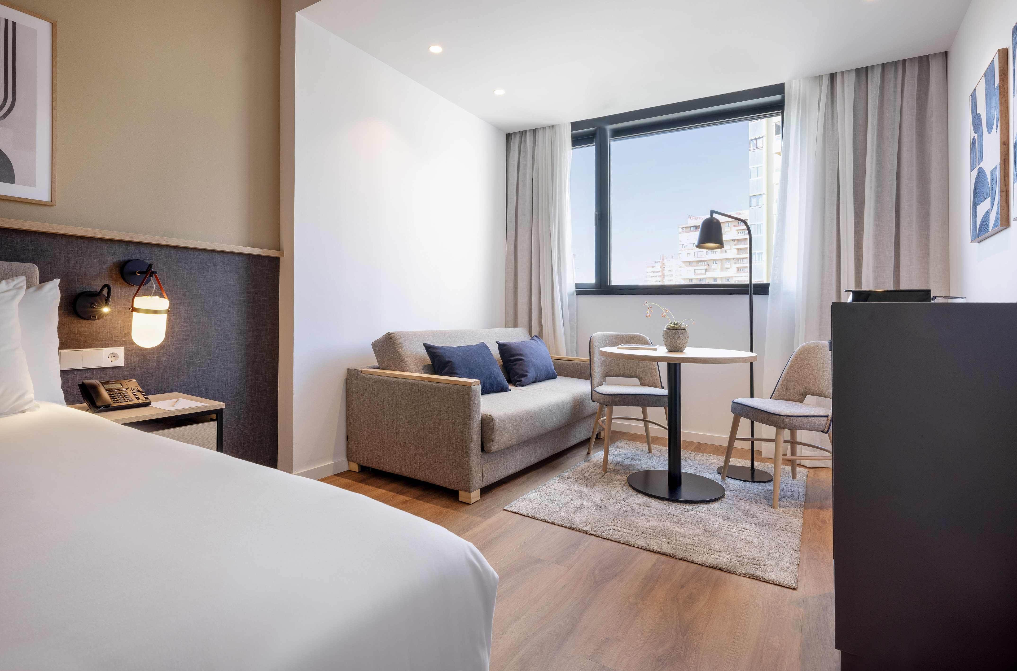 Modern hotel studio with bed, sofa, dining table, and city view window.