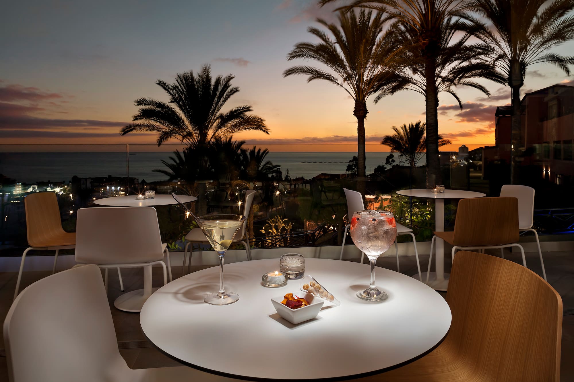 Cócteles en terraza con vista al atardecer sobre el mar y palmeras.