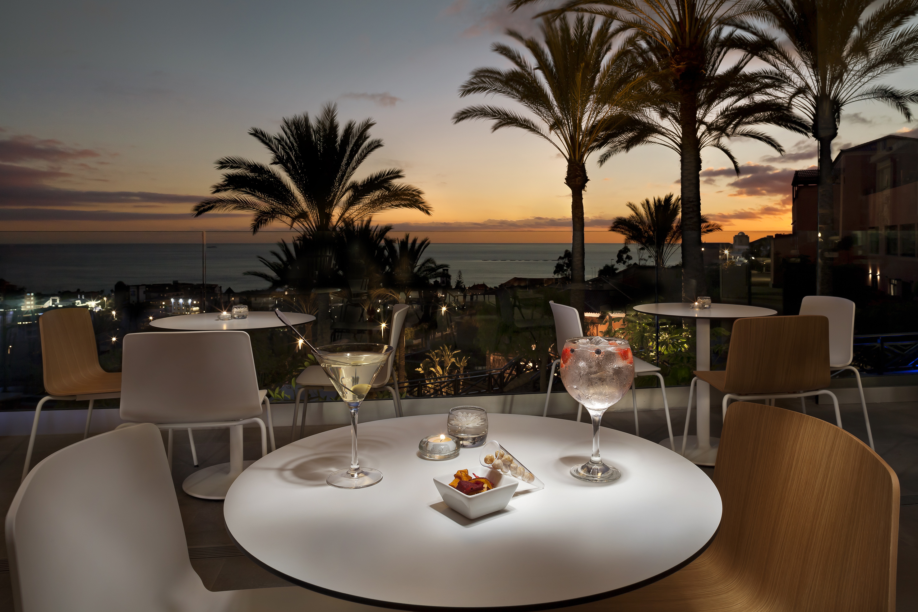 Cócteles en terraza con vista al atardecer sobre el mar y palmeras.