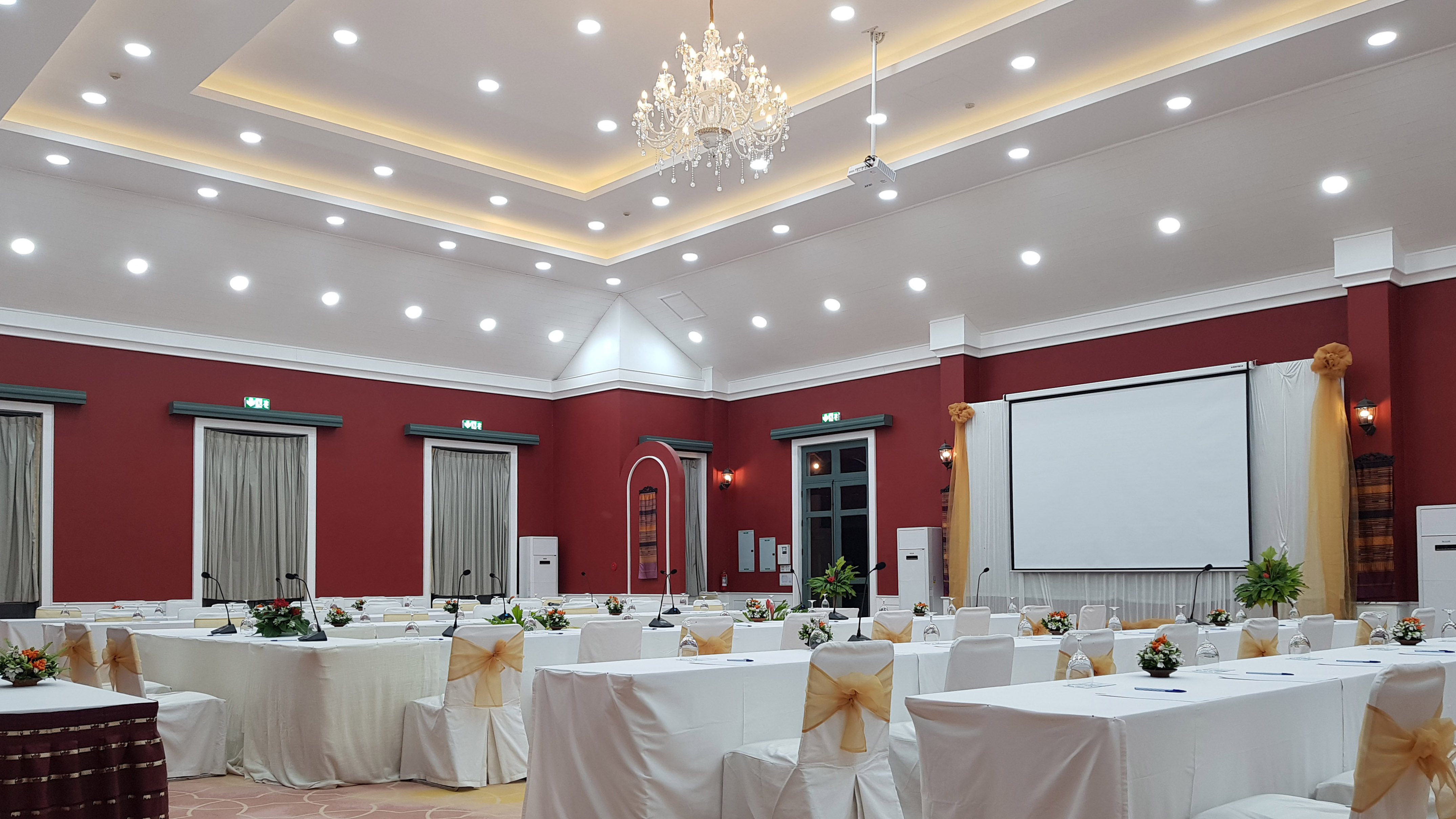 Elegant conference room with red walls, white tables, projector screen, and chandelier.