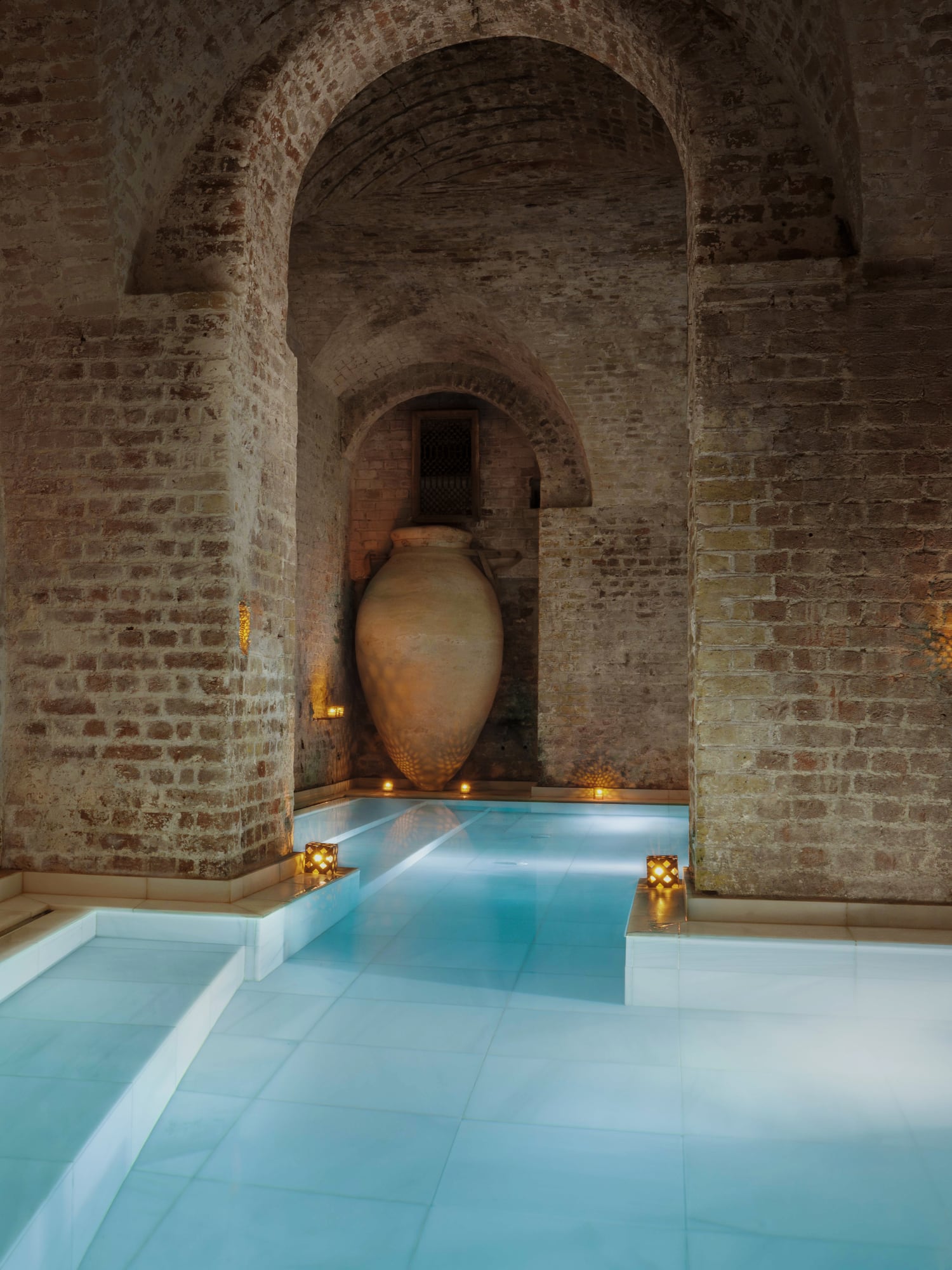 Arched brick Hamam spa with a large urn, glowing candles, and tranquil blue pool.