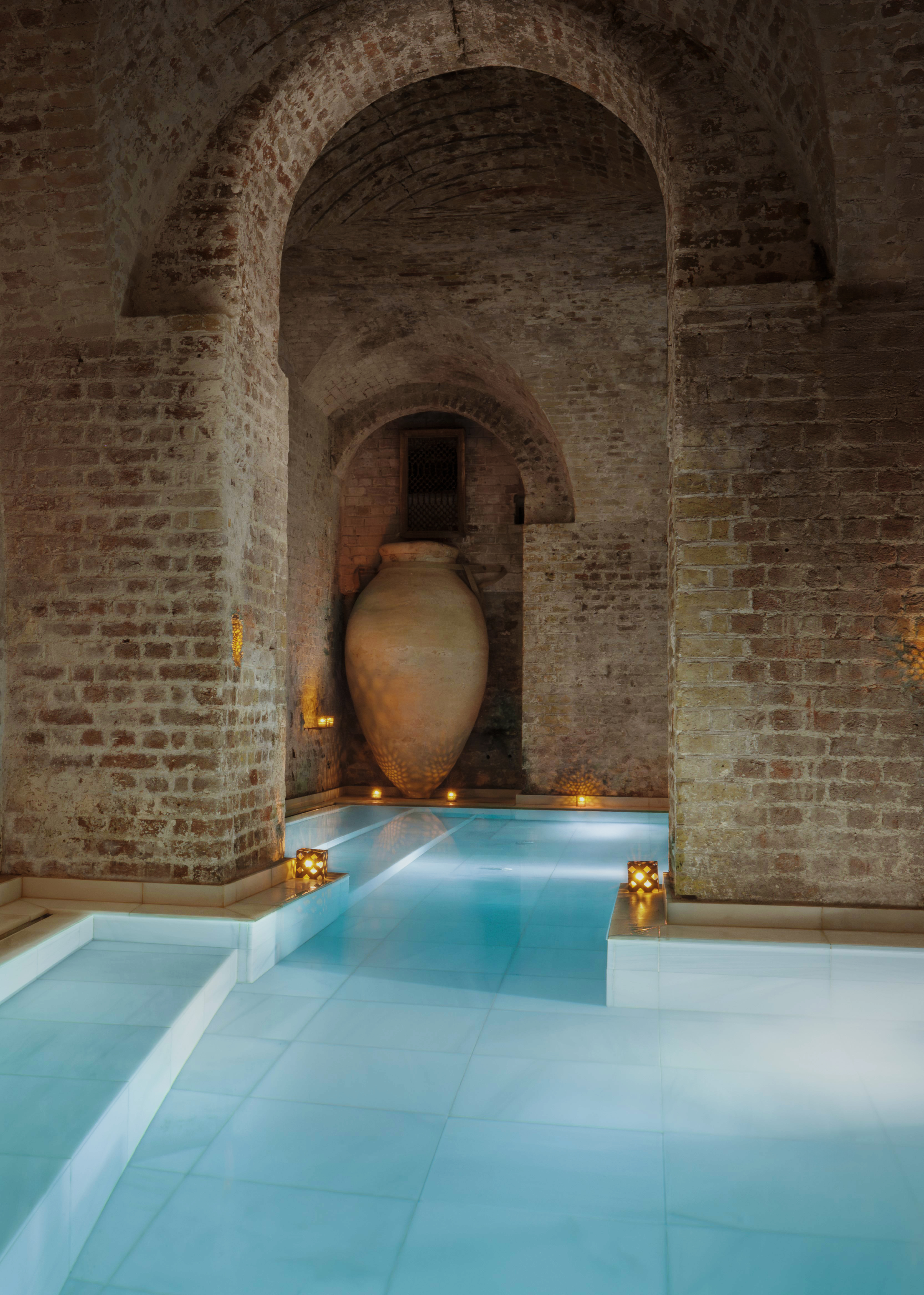Arched brick Hamam spa with a large urn, glowing candles, and tranquil blue pool.