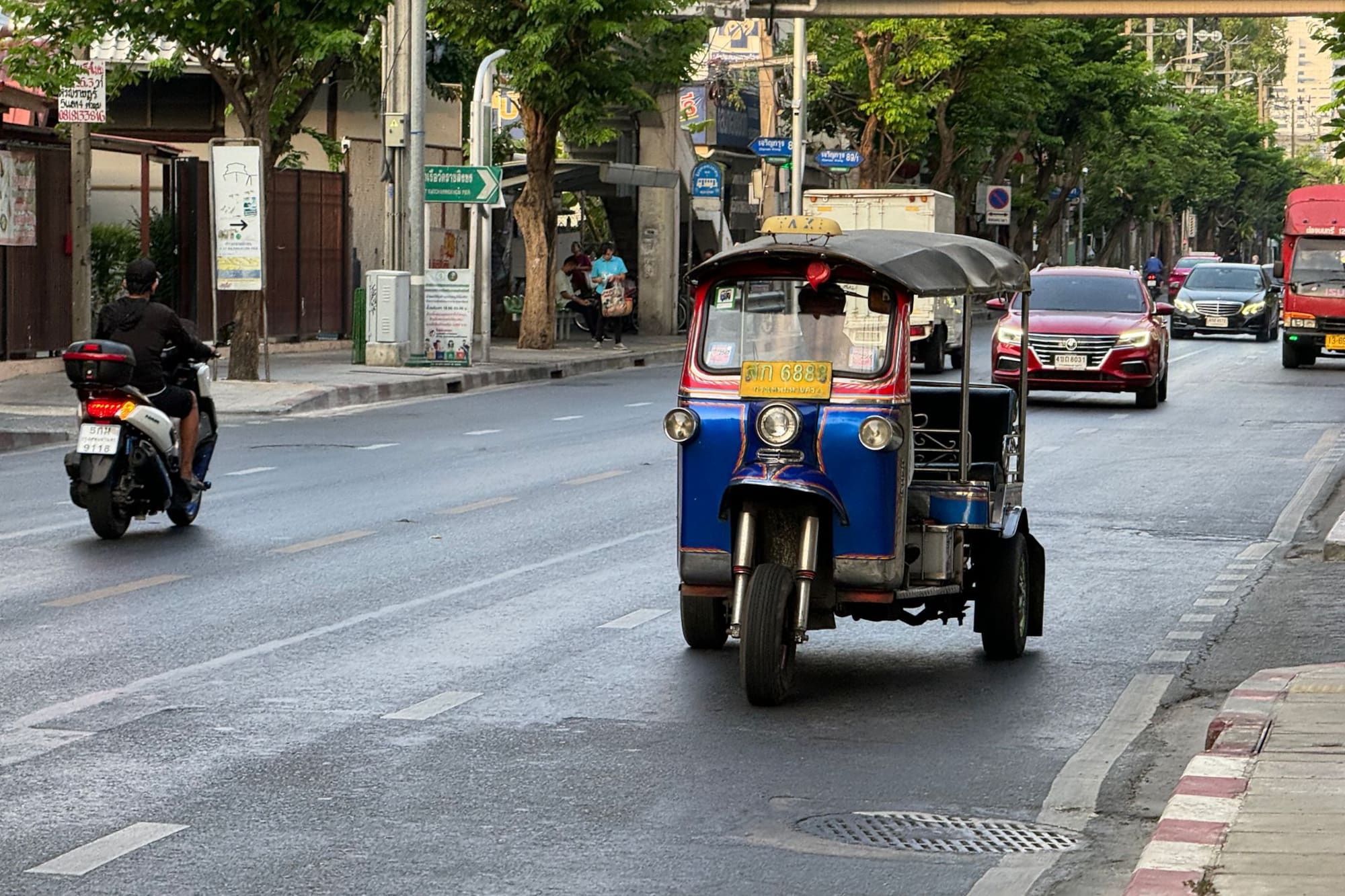 Blue and red tuk-tuk, motorcycle, and cars on a Bangkok city street.