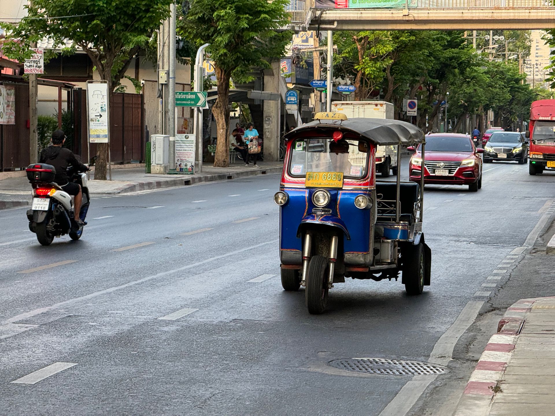 Blue and red tuk-tuk, motorcycle, and cars on a Bangkok city street.