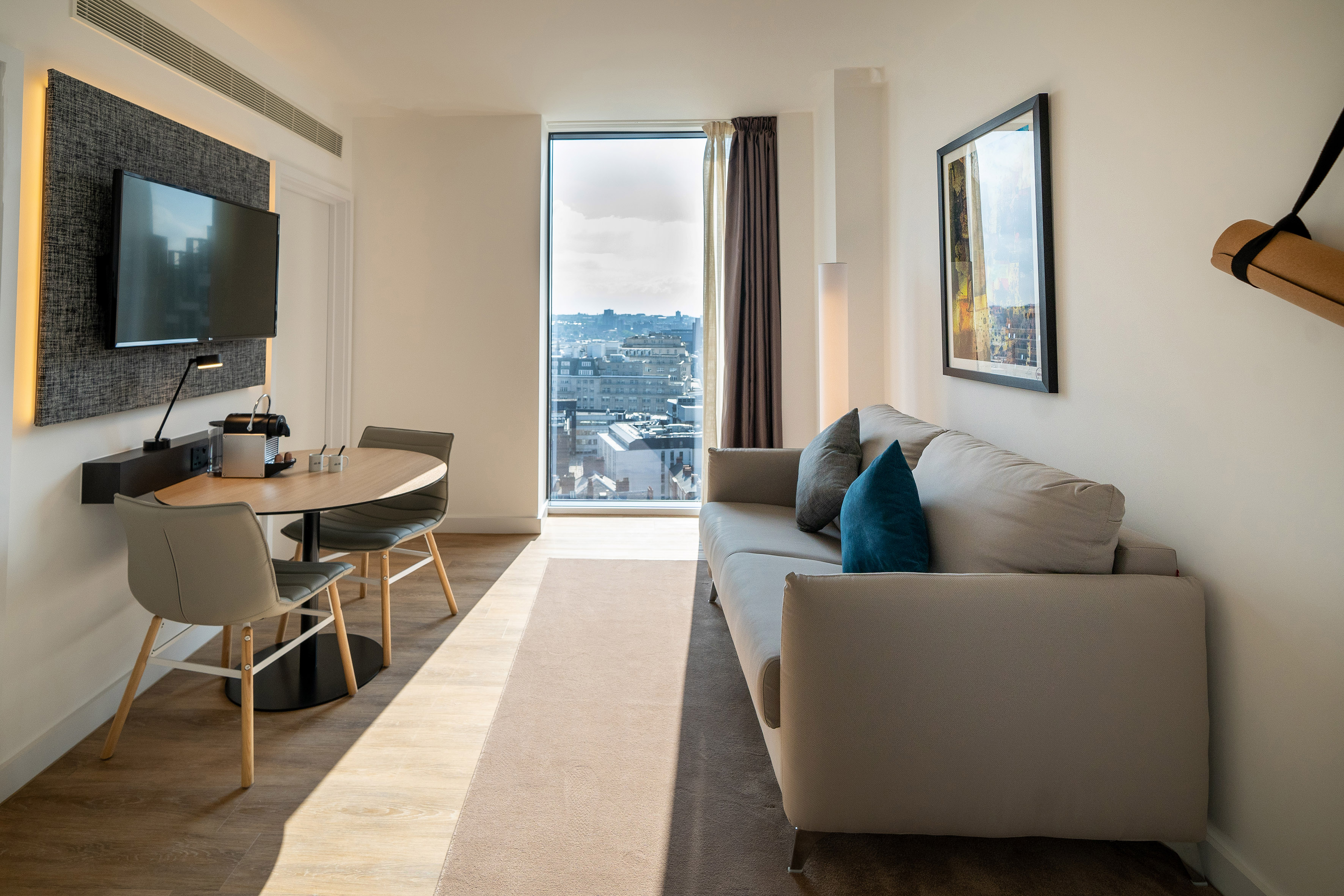Modern hotel living area with sofa, city view, dining table, and TV.