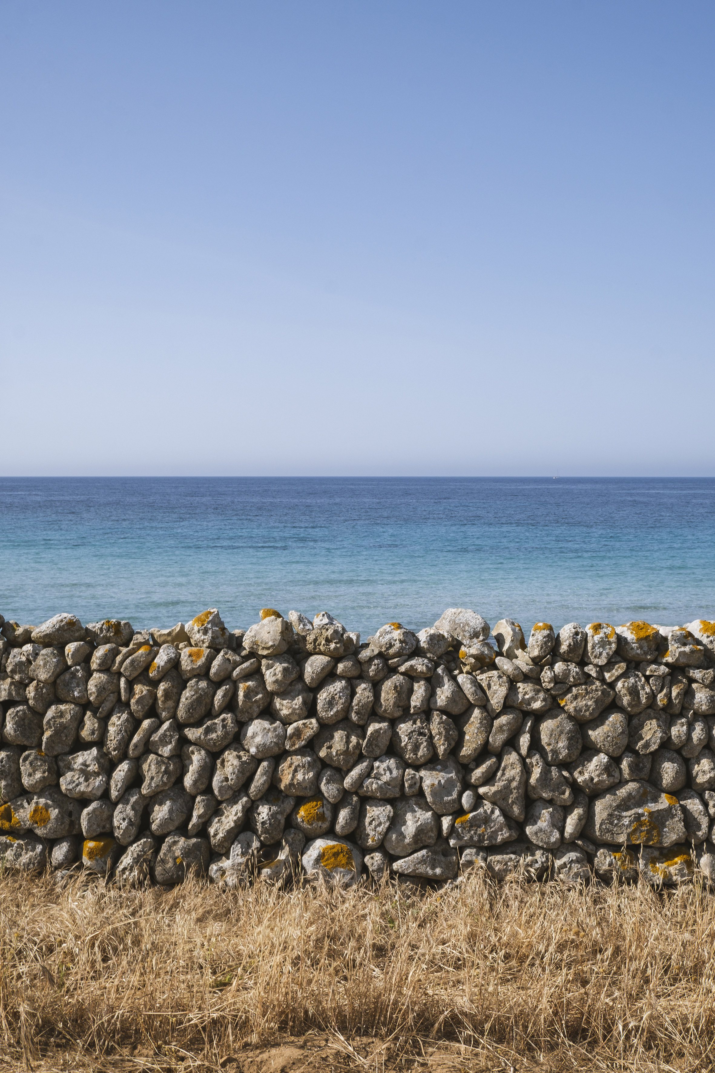 Paisaje costero: muro de piedra seca, hierba, mar azul y cielo.