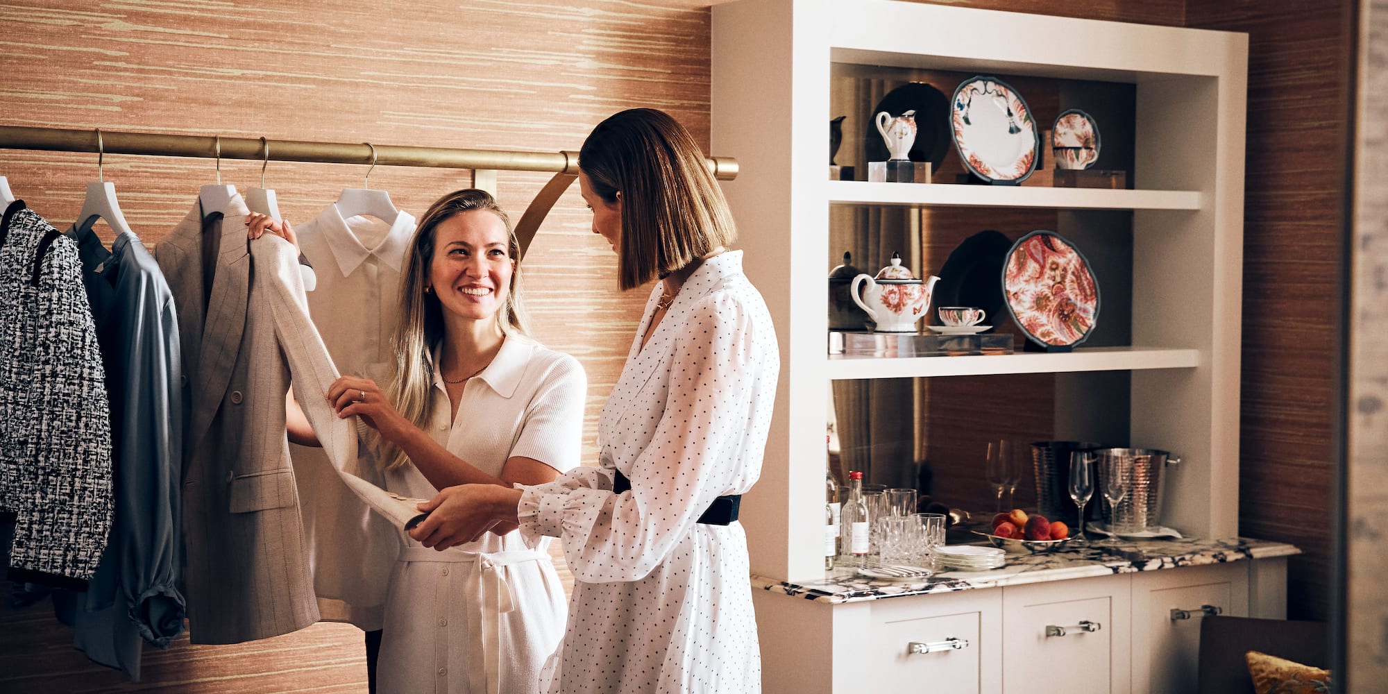 Women choosing blazer from clothing rack in stylish boutique or hotel room.