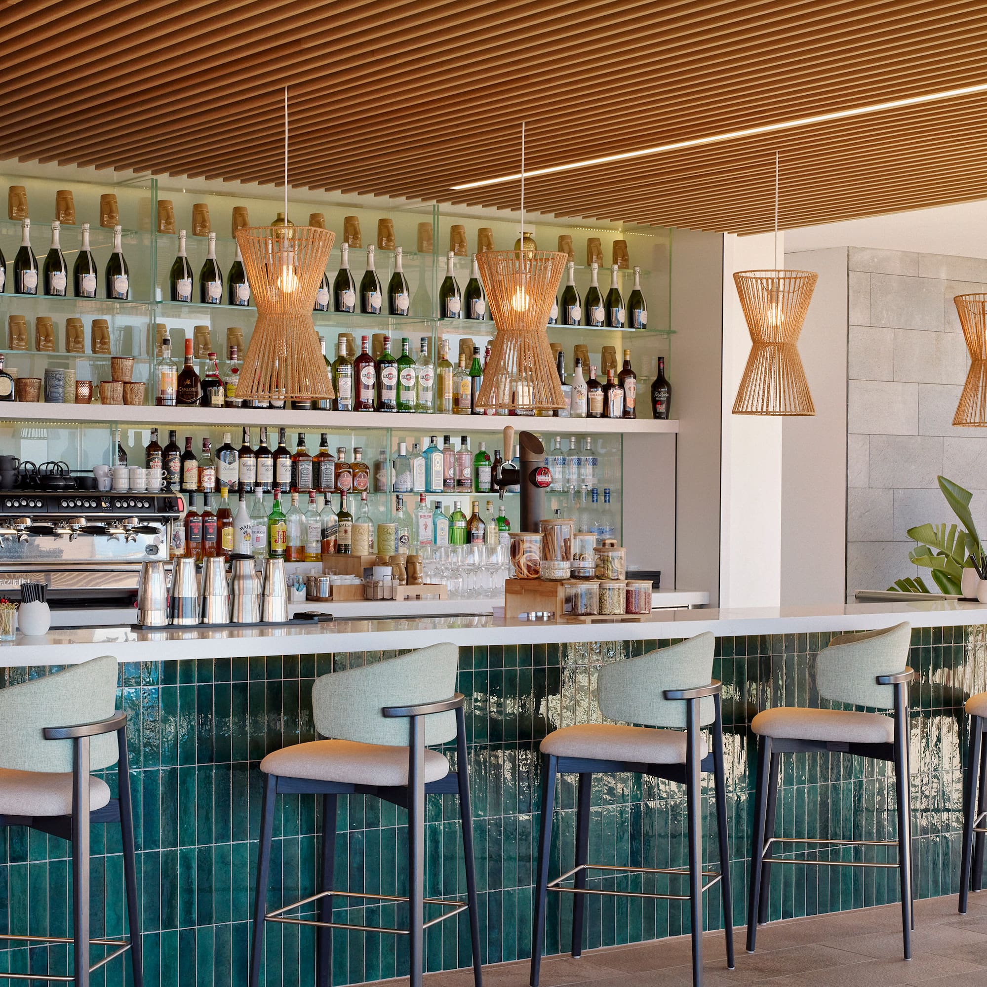 Modern bar with green tiled counter, rattan lights, and stocked beverage shelves.