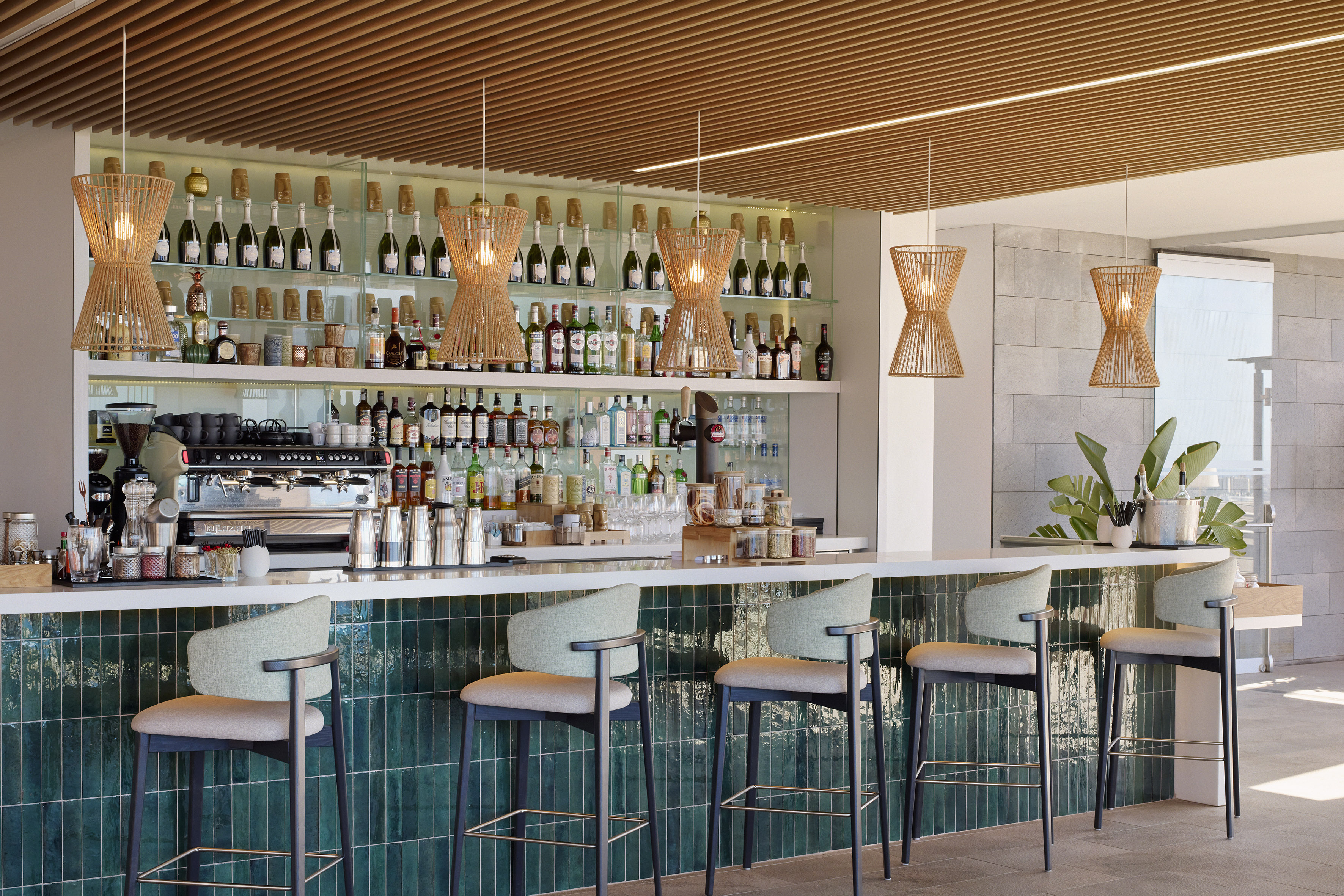Modern bar with green tiled counter, rattan lights, and stocked beverage shelves.