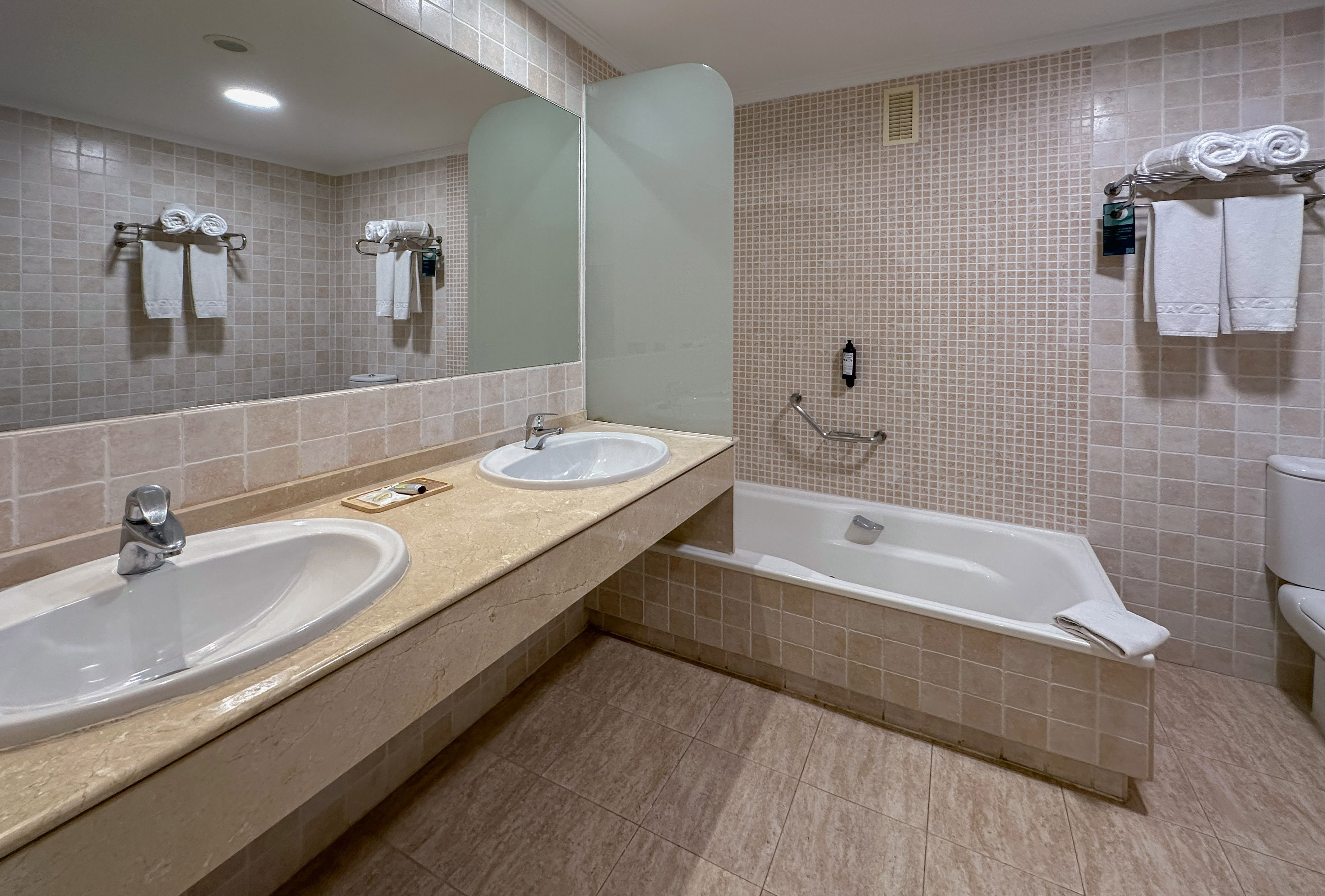 Beige tiled hotel bathroom with double sink vanity, mirror, and bathtub.