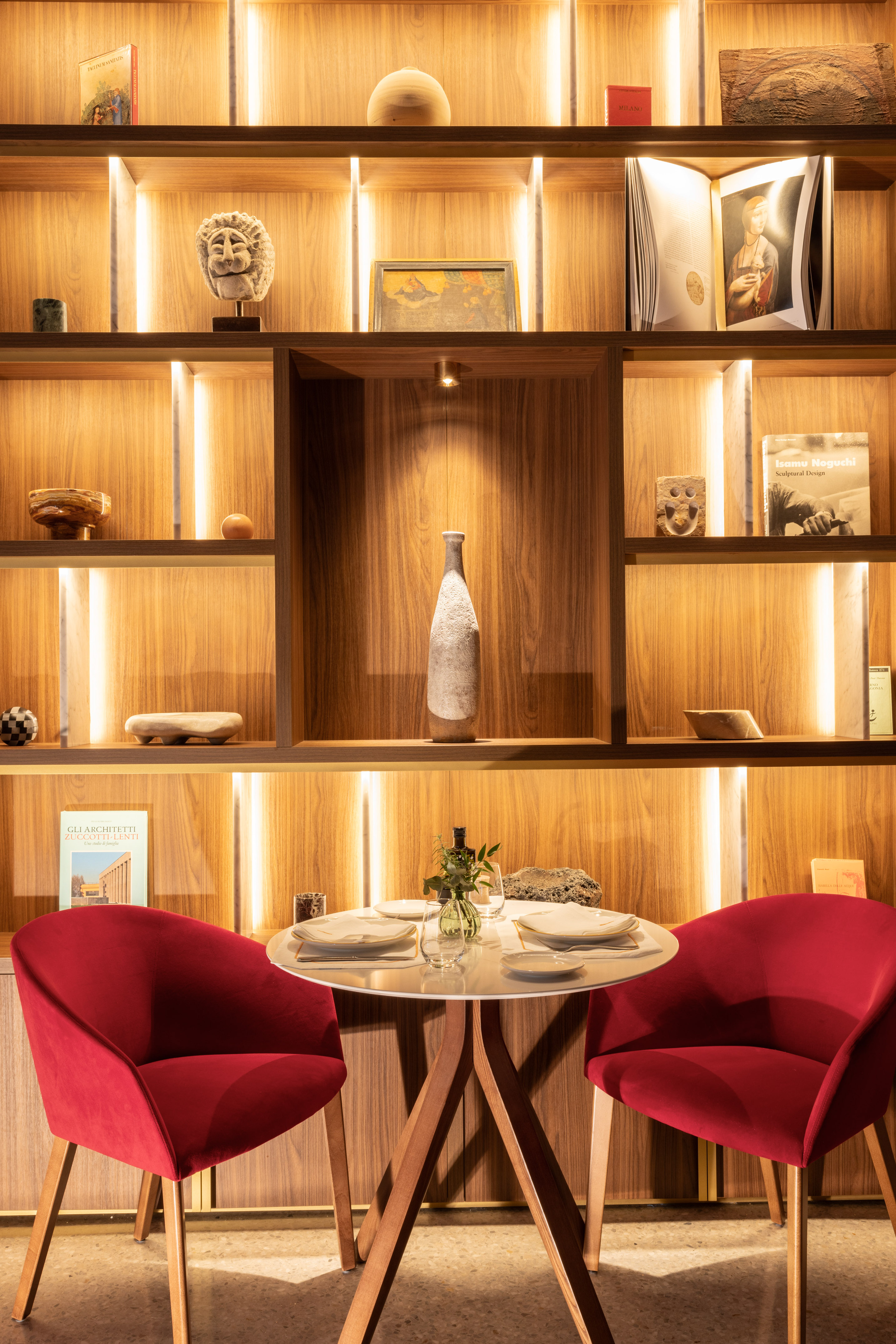 Restaurant dining area with illuminated wooden shelves, white table, red chairs.