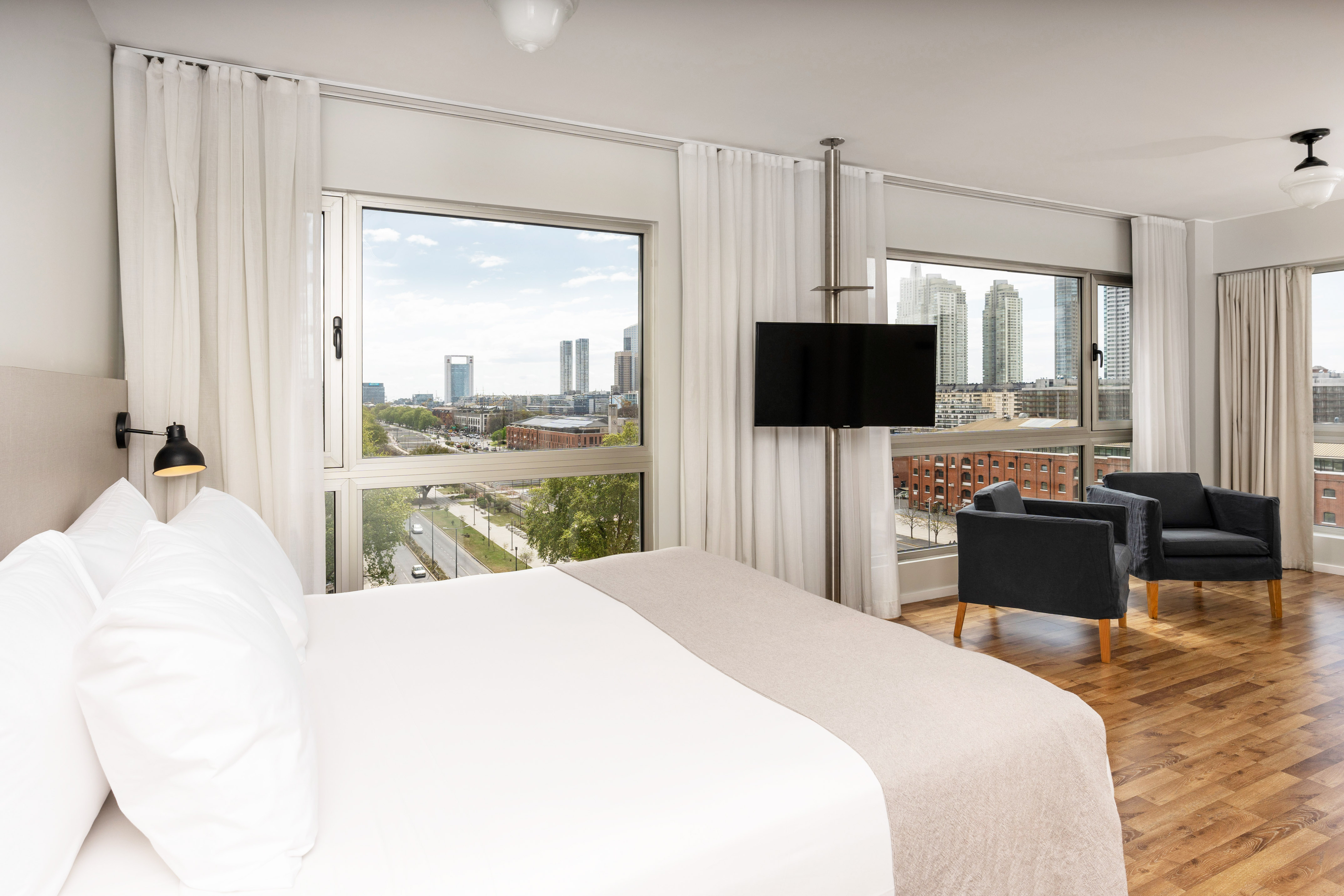Hotel bedroom with city view, white bed, TV, and two armchairs.