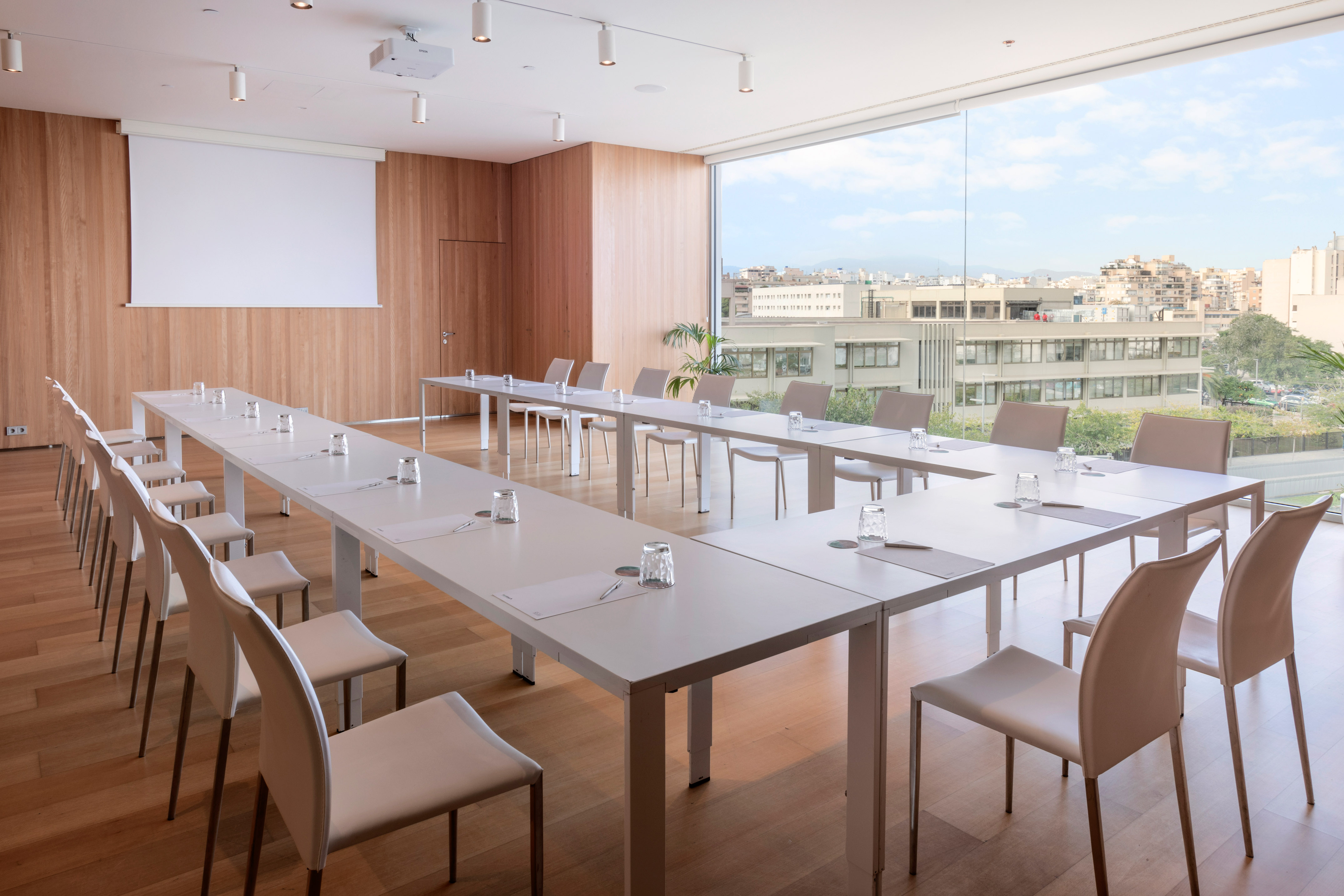 Modern meeting room with U-shaped tables, projector, city view, wood accents.
