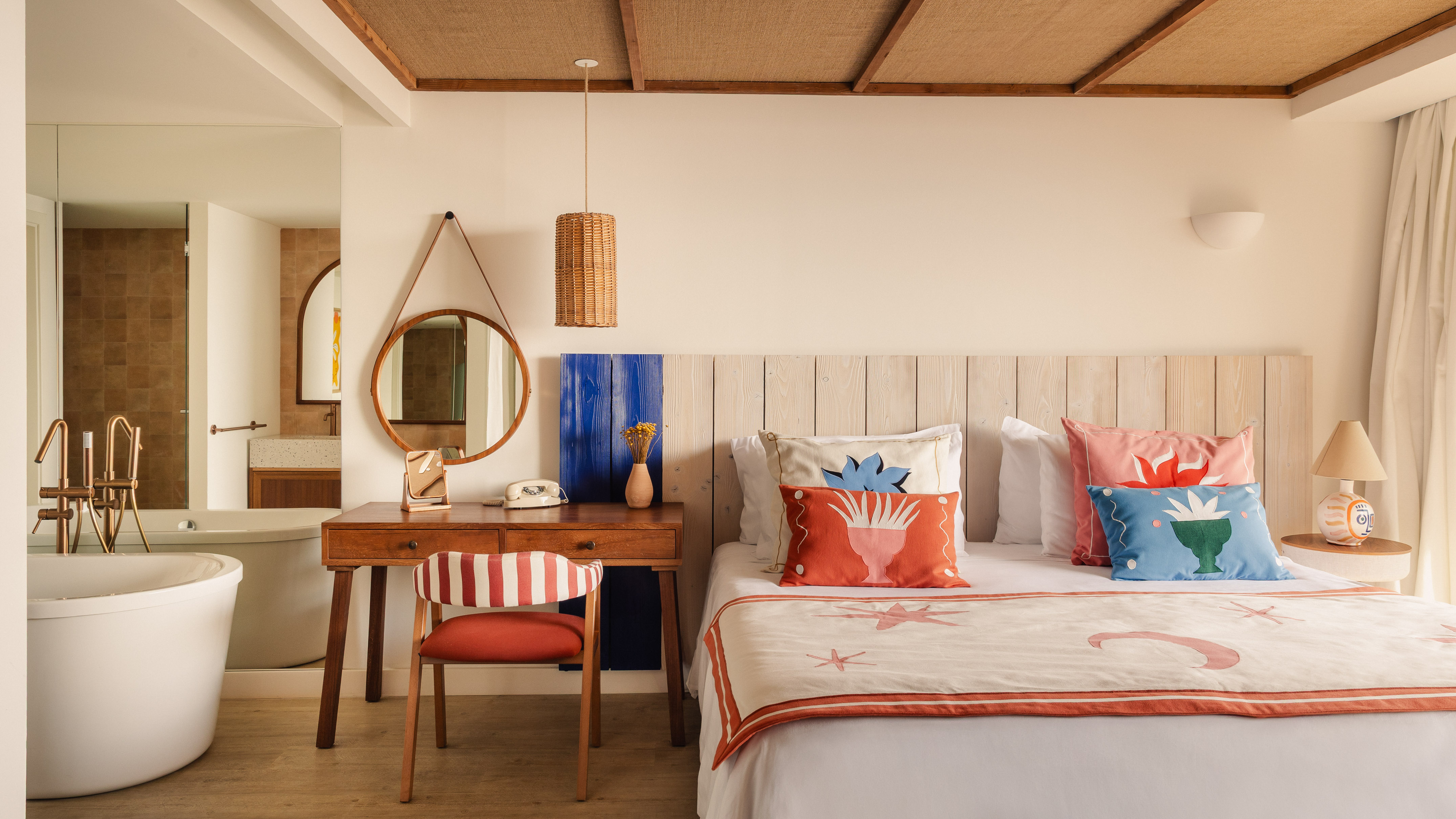 Bright hotel bedroom with colorful bed, wooden desk, round mirror, and open bathroom.