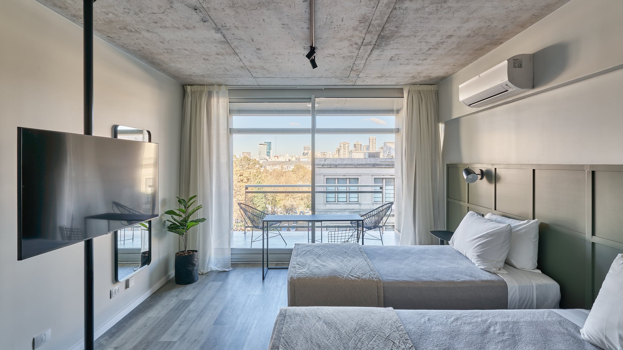 Modern twin bed hotel room with city view balcony and concrete ceiling.