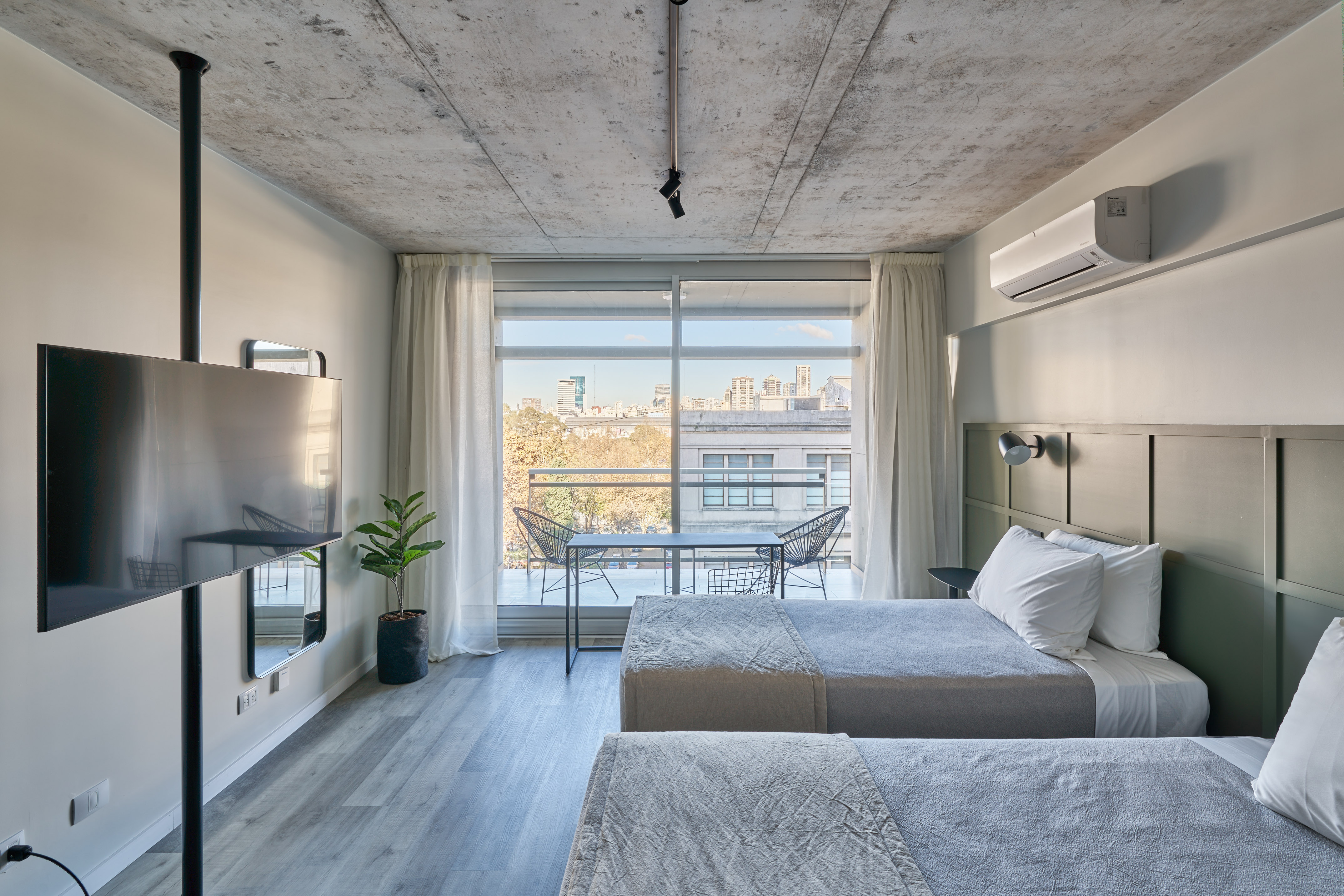Modern twin bed hotel room with city view balcony and concrete ceiling.