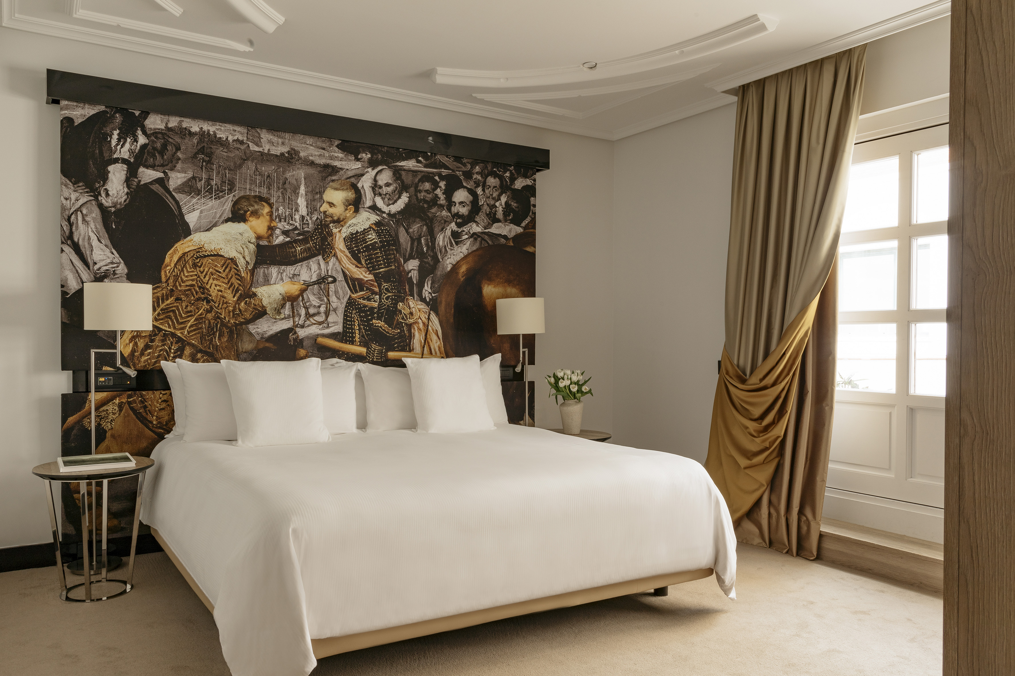 Luxury hotel bedroom with large historic art mural above king bed and gold curtains.