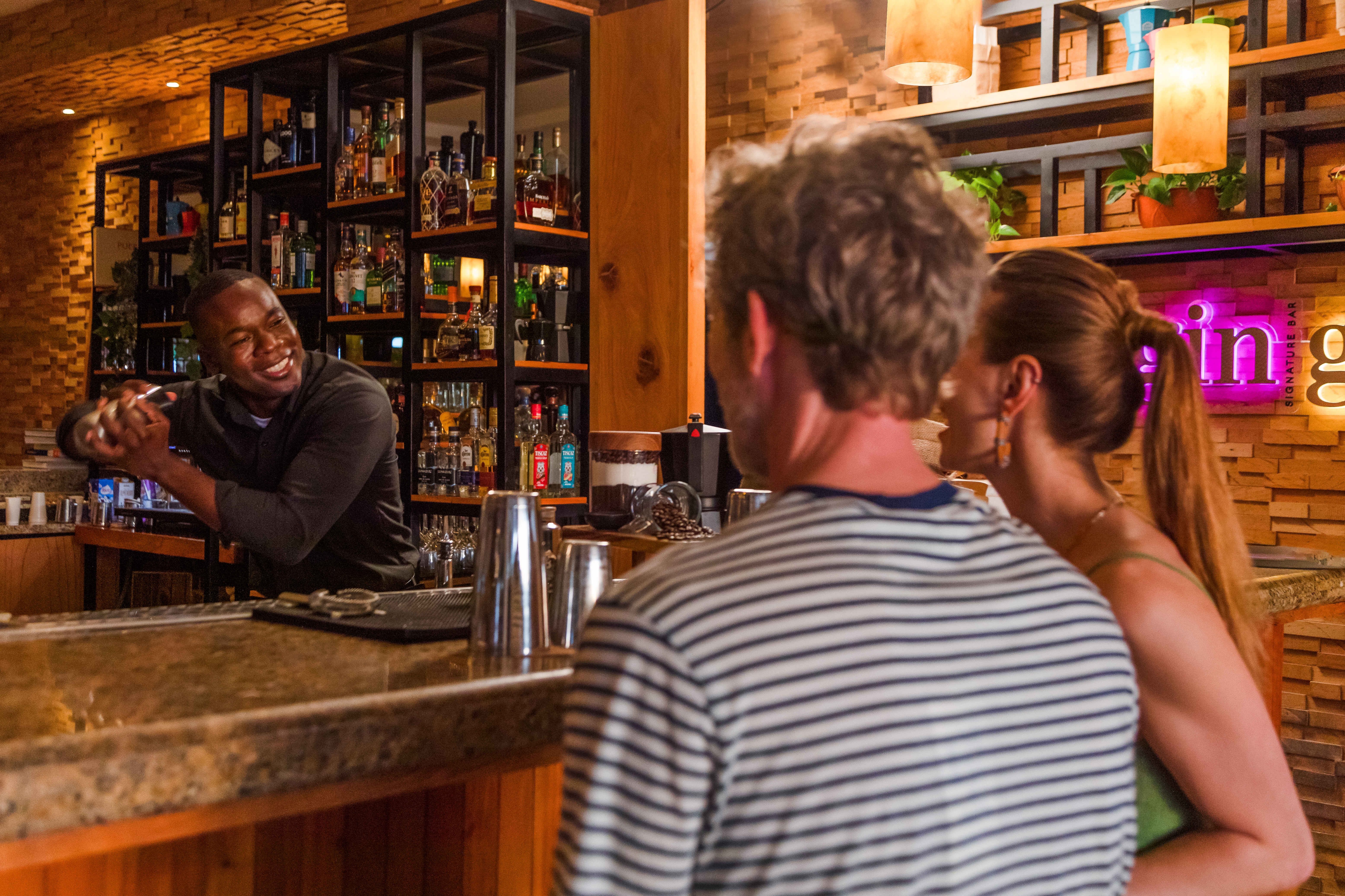 Smiling bartender shaking cocktail for customers at a lively bar with liquor bottles.
