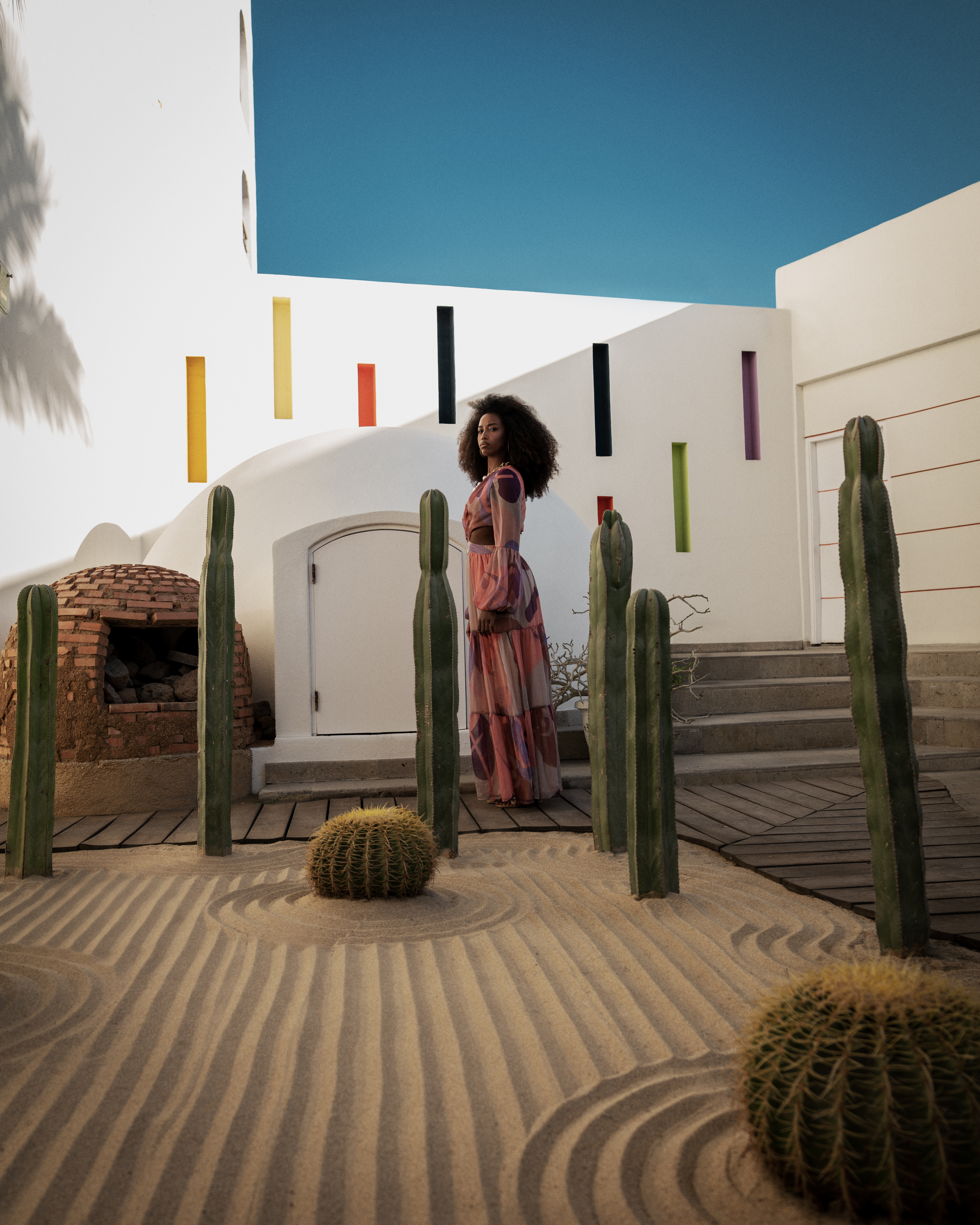 Woman in patterned dress, desert zen garden with cacti, white modern building.