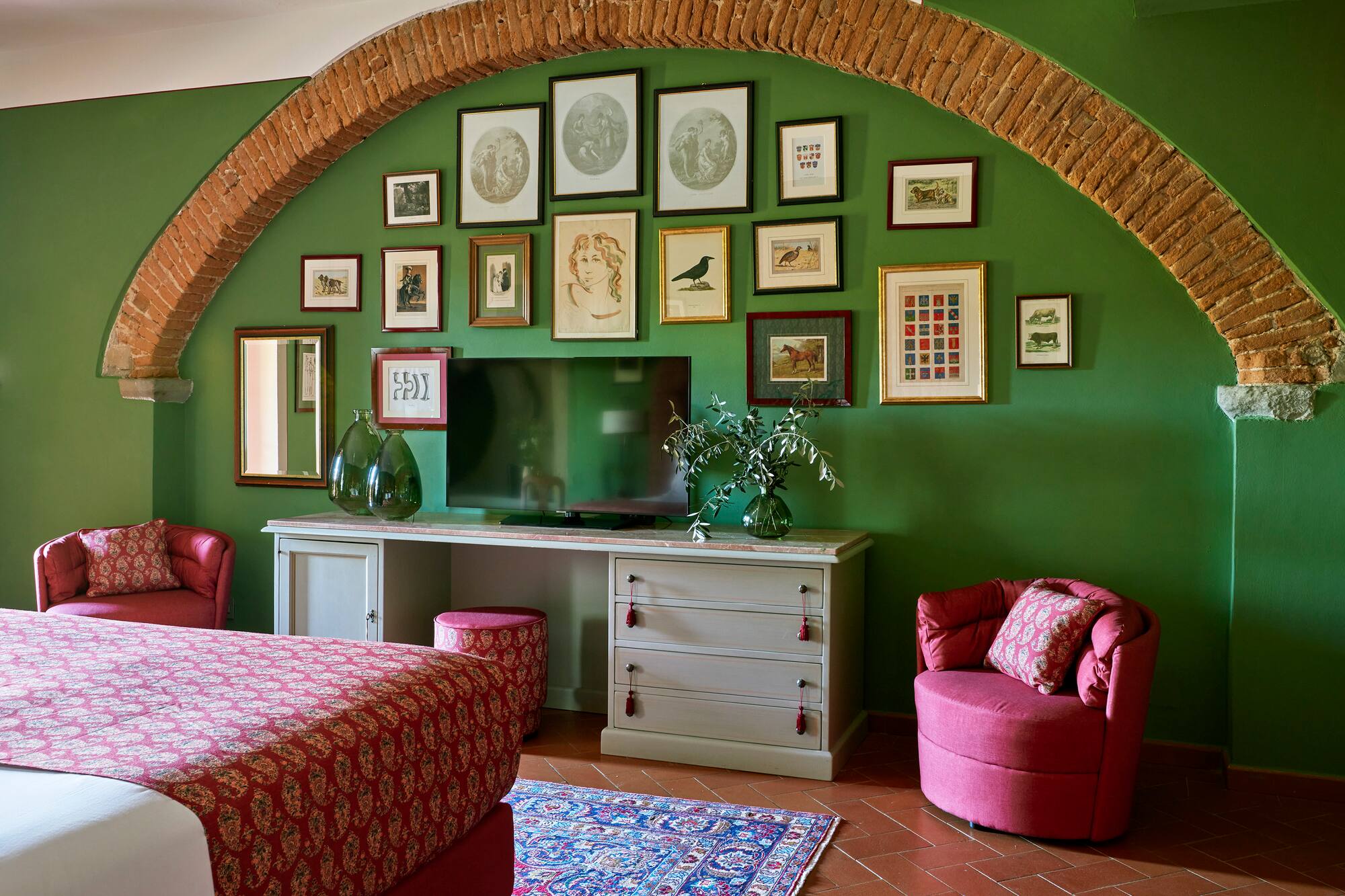 Green bedroom with brick arch, art gallery wall, red bed, and terracotta tiles.