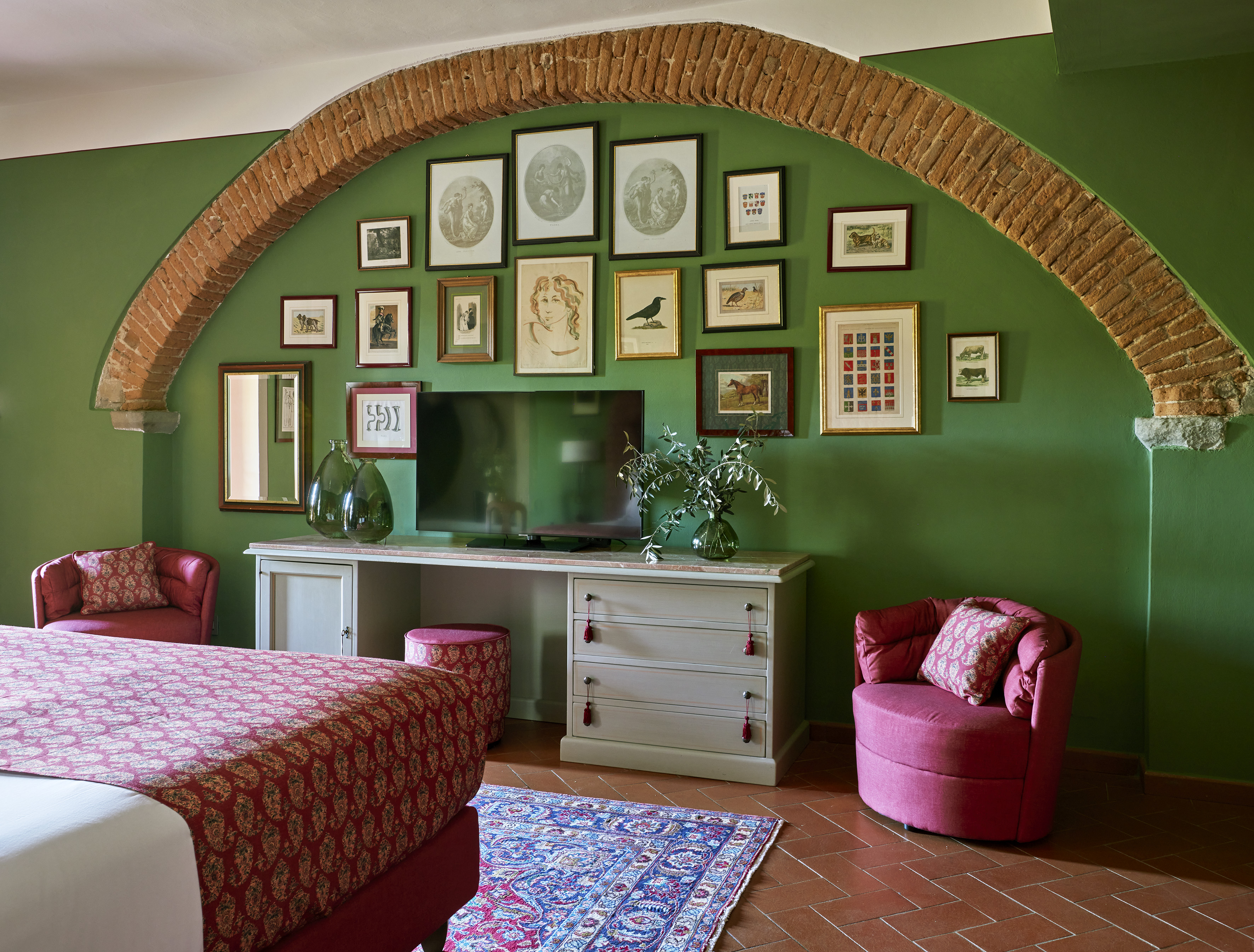 Green bedroom with brick arch, art gallery wall, red bed, and terracotta tiles.