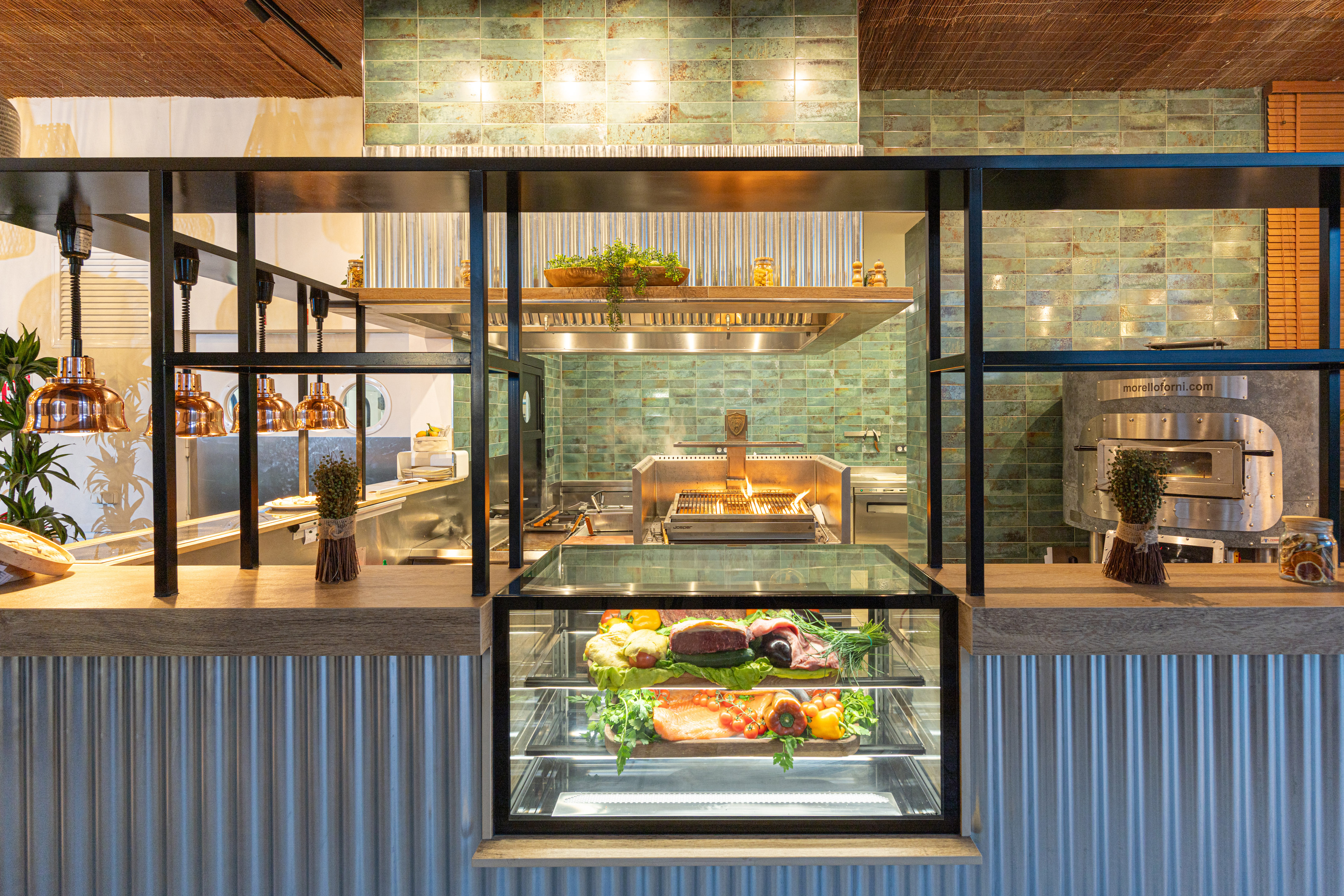 Modern open kitchen with grill, fresh meat and produce display, and pizza oven.