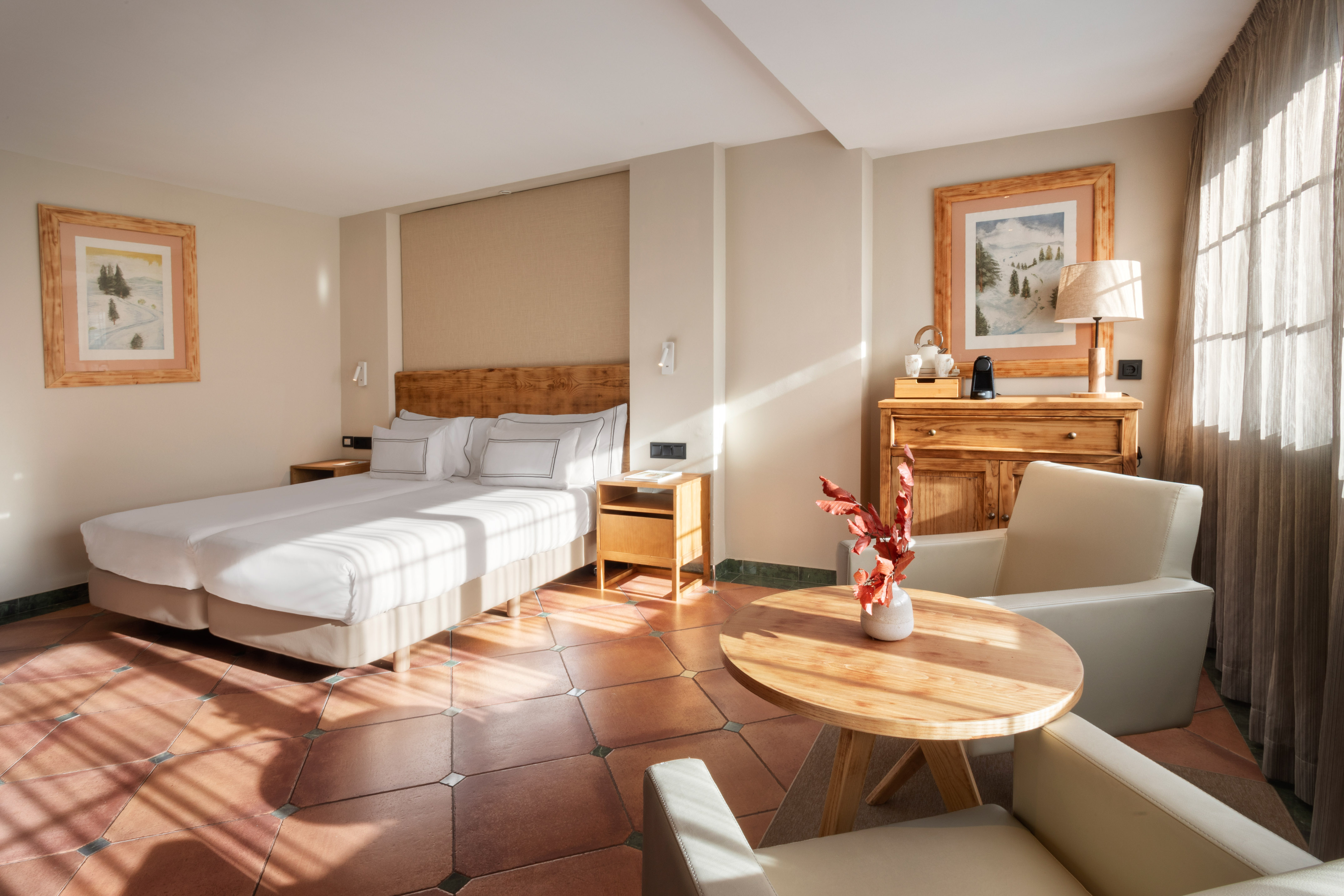 Sunlit cozy hotel bedroom with two beds, wooden furniture, and terracotta floor.