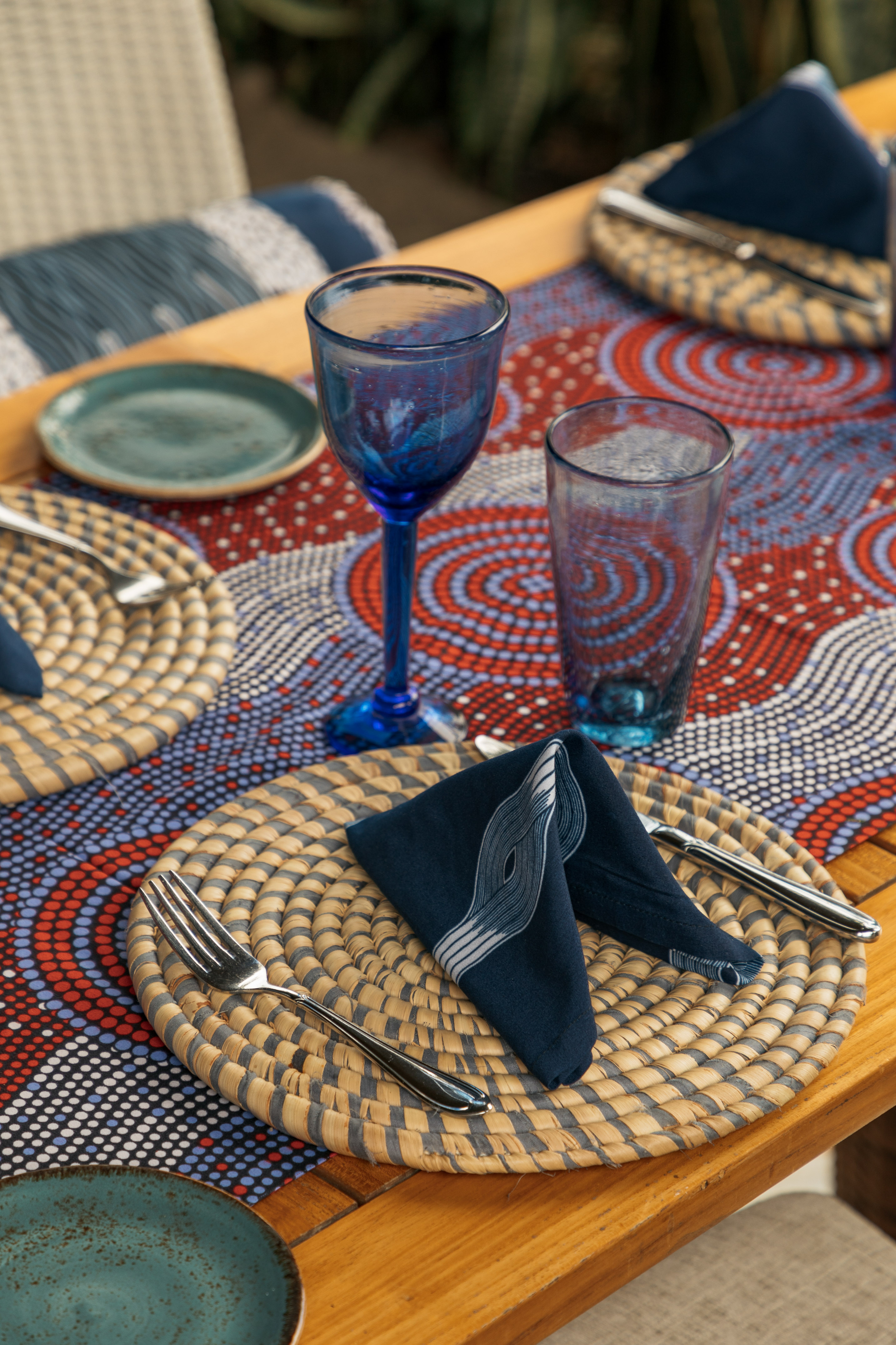 Vibrant outdoor table setting with woven placemats, blue glassware, patterned runner.