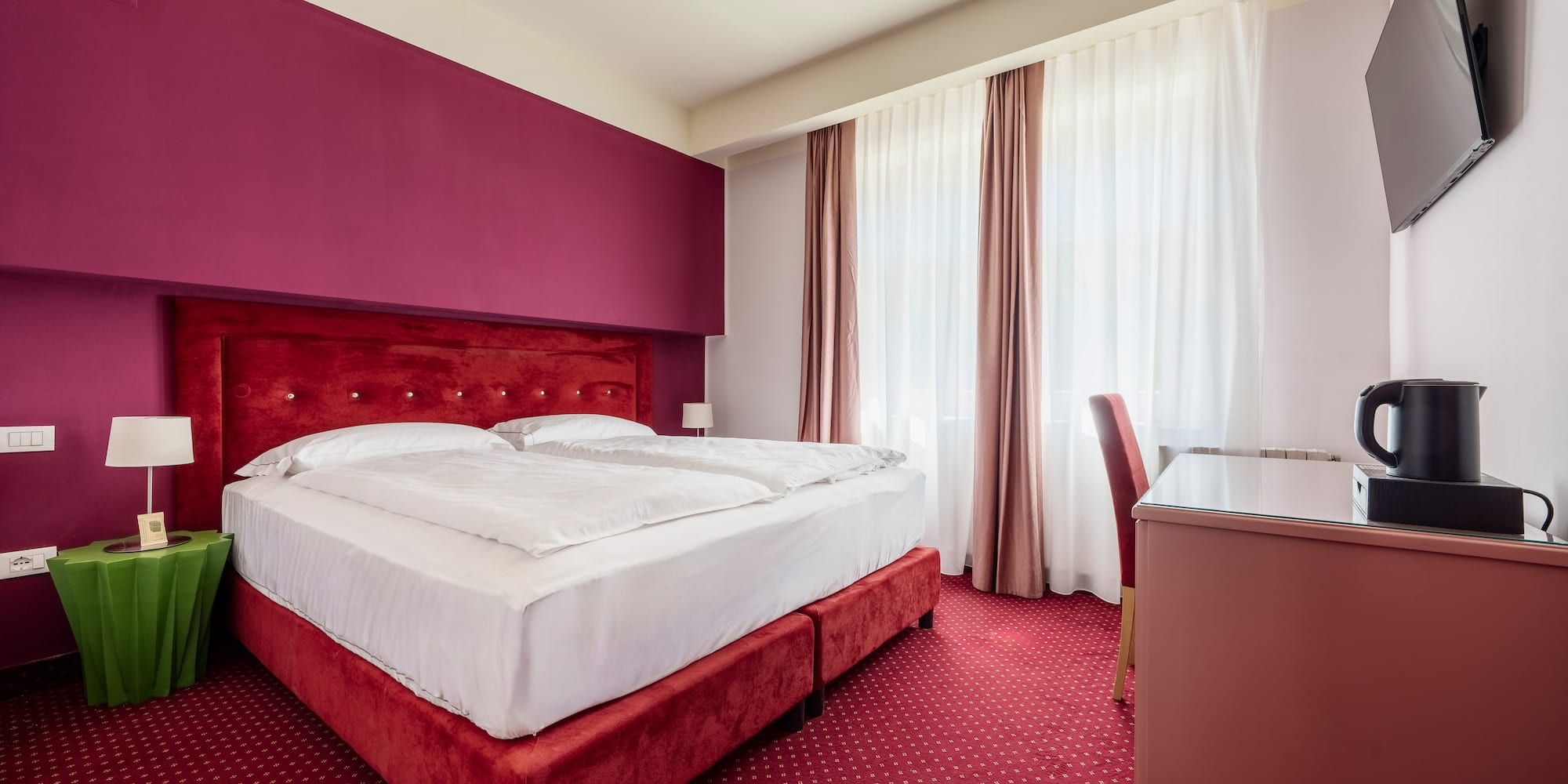 Vibrant hotel room with red double bed, fuchsia accent wall, and patterned carpet.