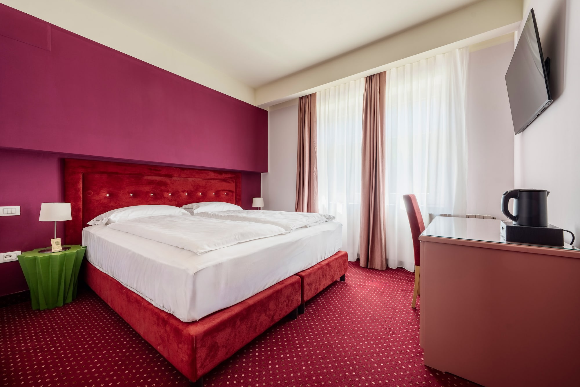 Vibrant hotel room with red double bed, fuchsia accent wall, and patterned carpet.