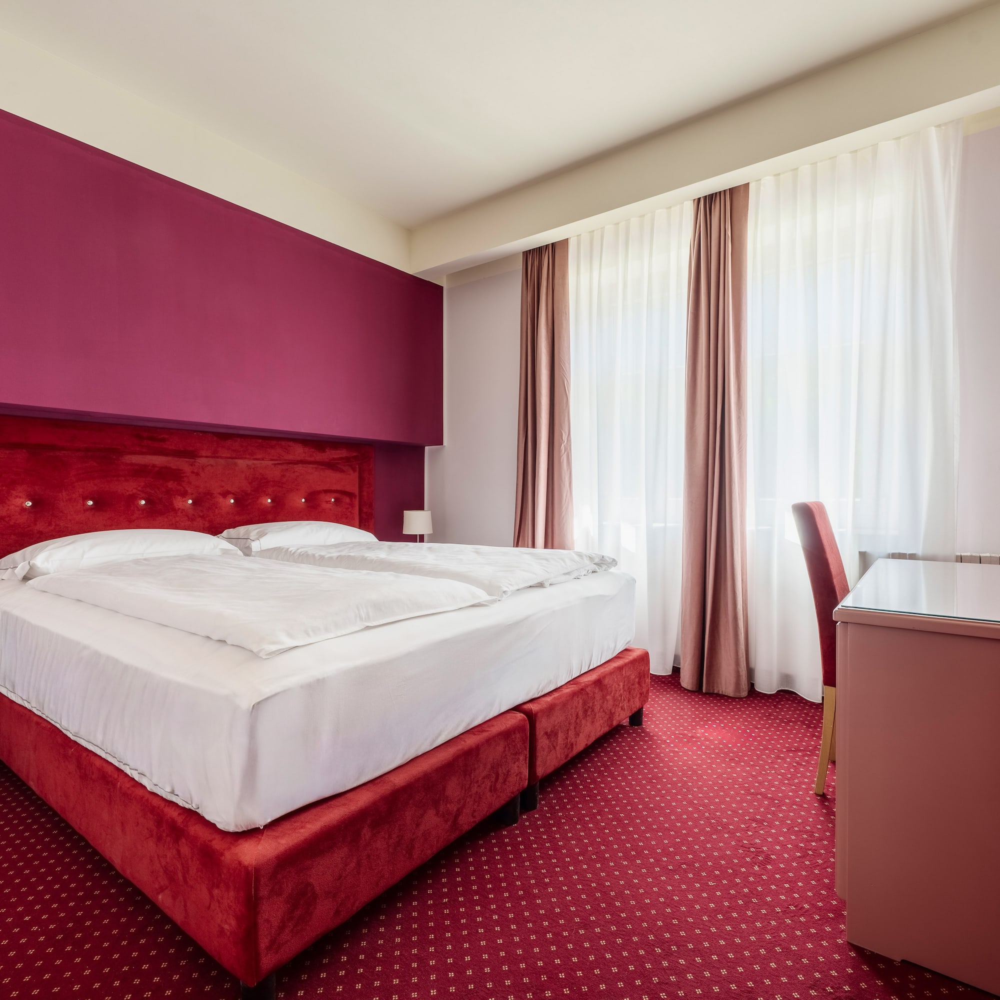 Vibrant hotel room with red double bed, fuchsia accent wall, and patterned carpet.