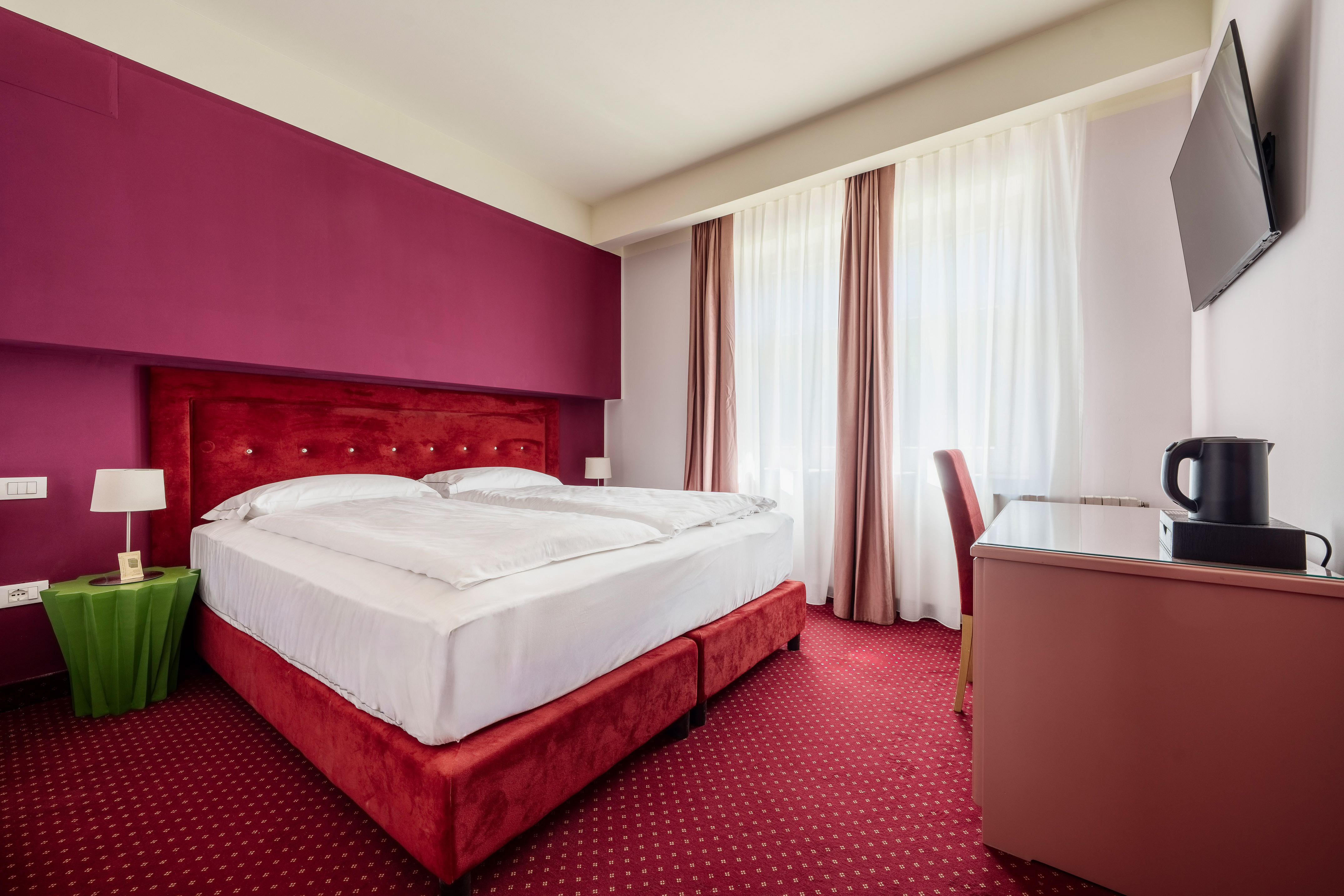Vibrant hotel room with red double bed, fuchsia accent wall, and patterned carpet.
