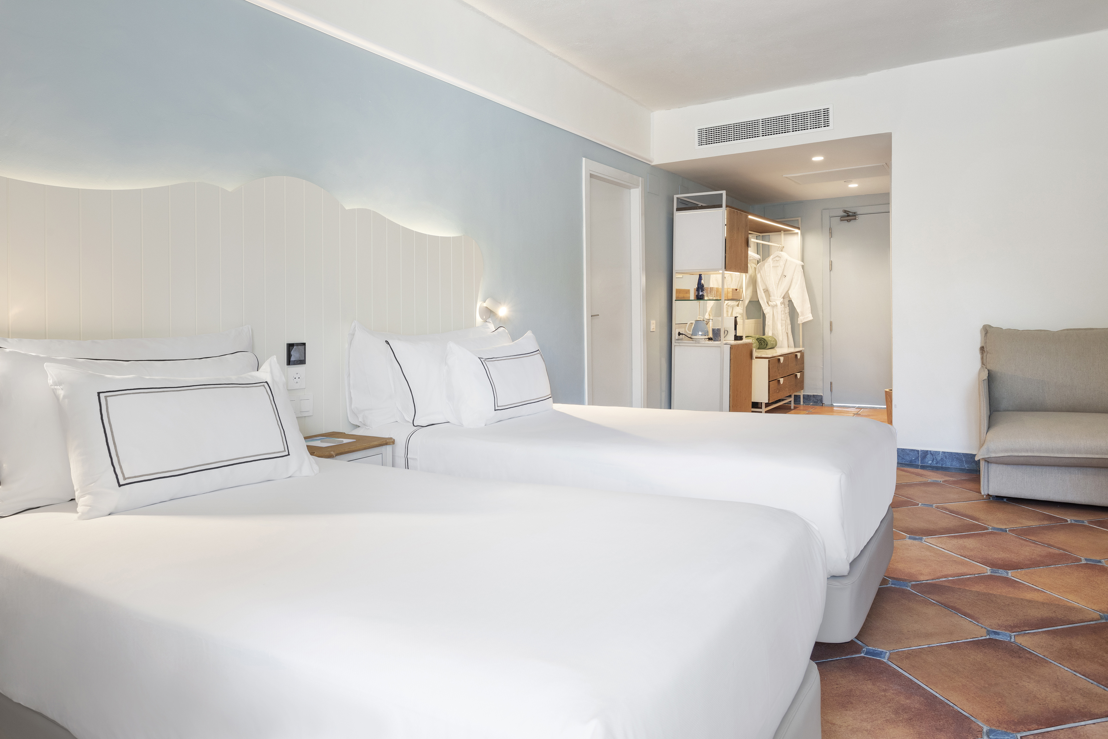 Modern hotel room with two twin beds, light blue wall, and terracotta floor.