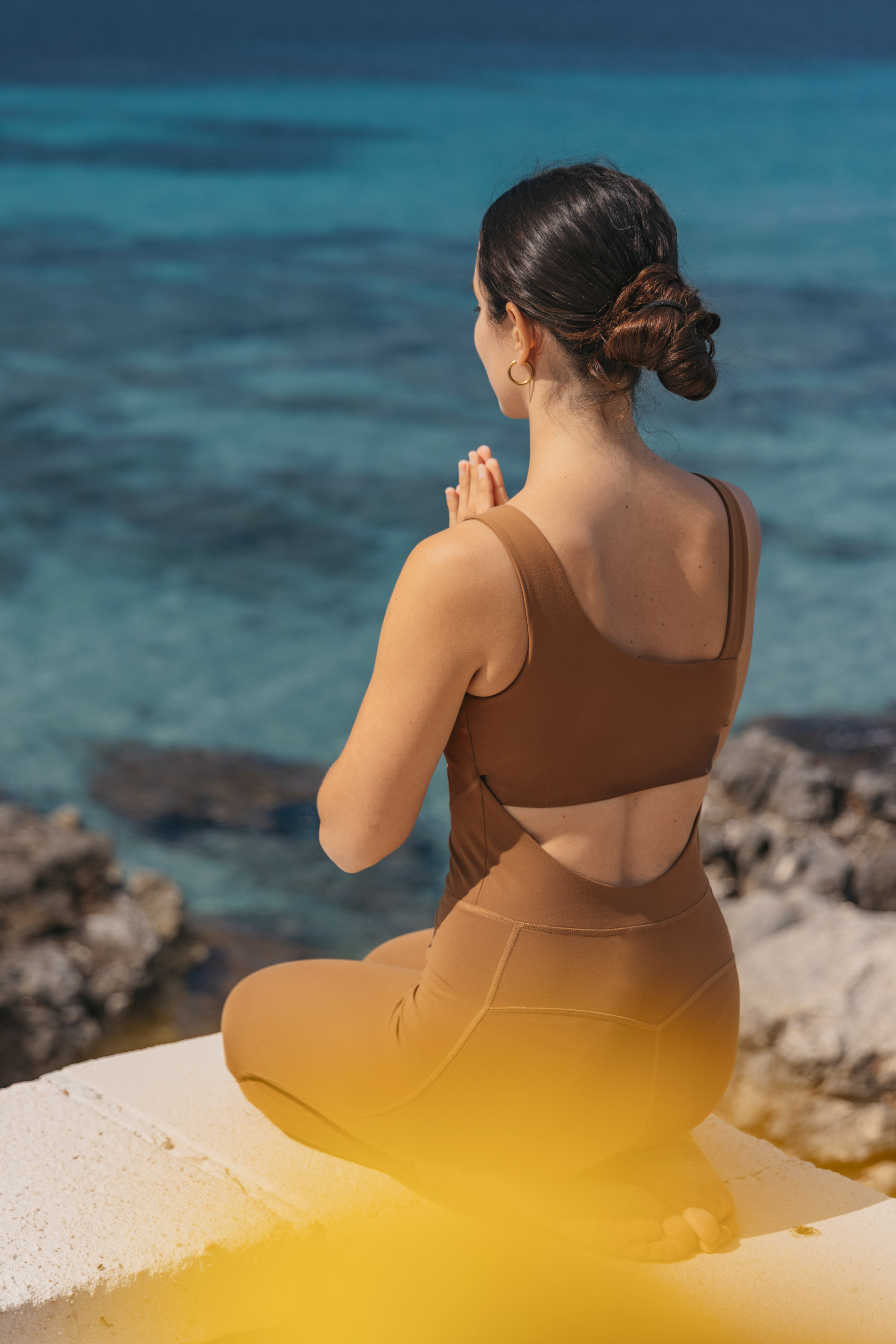 Woman meditating in brown activewear by blue ocean, hands in prayer.